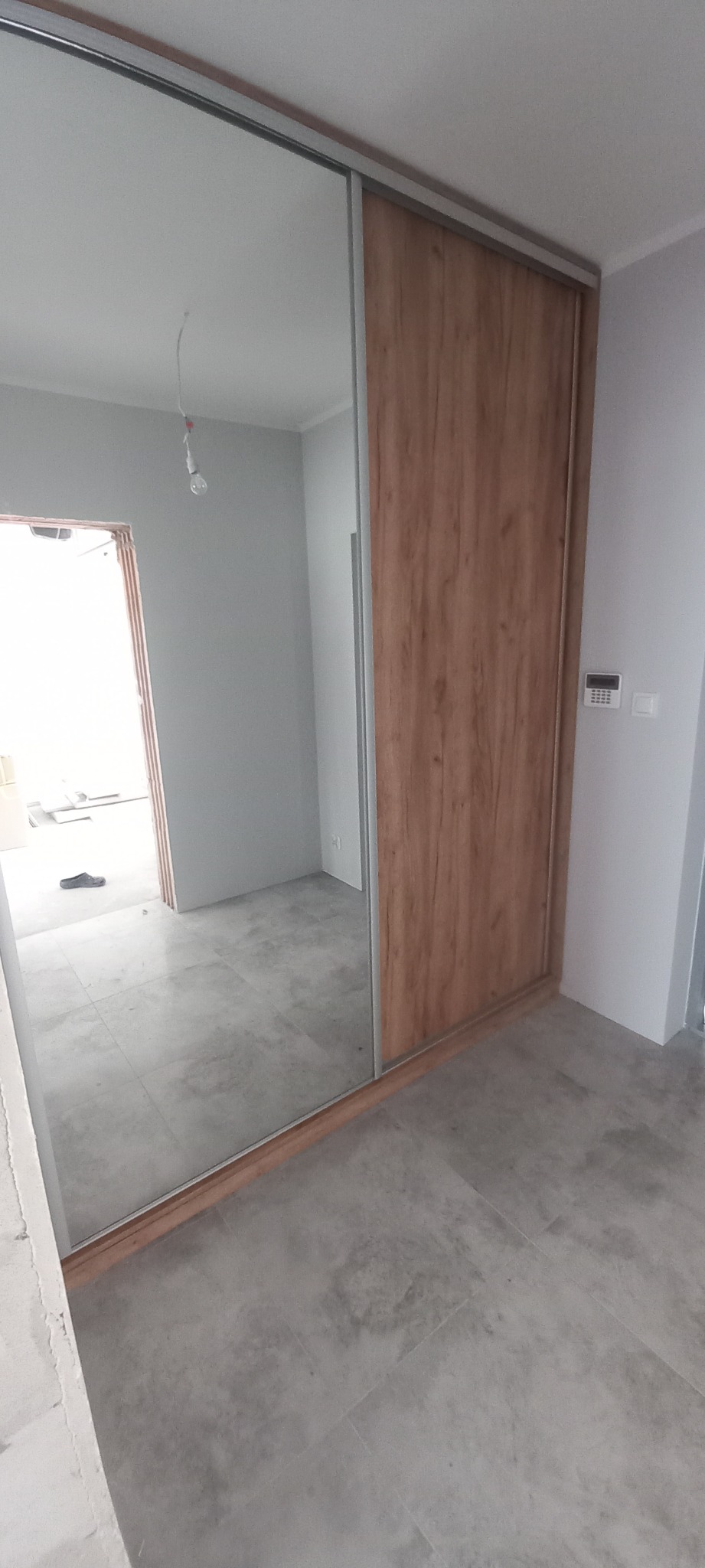Nowoczesna szafa wnękowa z przesuwnymi drzwiami: jedne lustrzane, drugie z okleiną imitującą drewno. Widoczna surowa podłoga z szarych płytek i niedokończone wykończenie ścian.