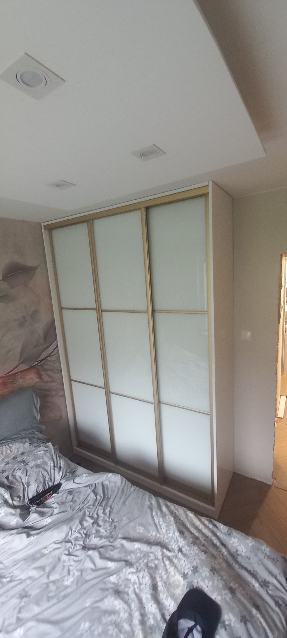 Wnętrze sypialni z białą szafą przesuwną z trzema matowymi szklanymi panelami w jasnych drewnianych ramach, widoczna część łóżka z szarą pościelą w kwiaty, na ścianie fototapeta z motywem roślinnym...