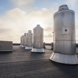 Turdach Szczecinek - Szereg metalowych kominów wentylacyjnych na płaskim dachu pokrytym papą, z widocznym żurawiem budowlanym w oddali i błękitnym niebem z chmurami.