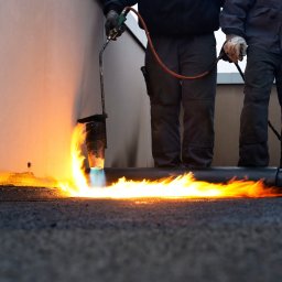 Turdach Szczecinek - Pracownik w kombinezonie spawa papę na dachu płomieniem palnika na tle ściany budynku.