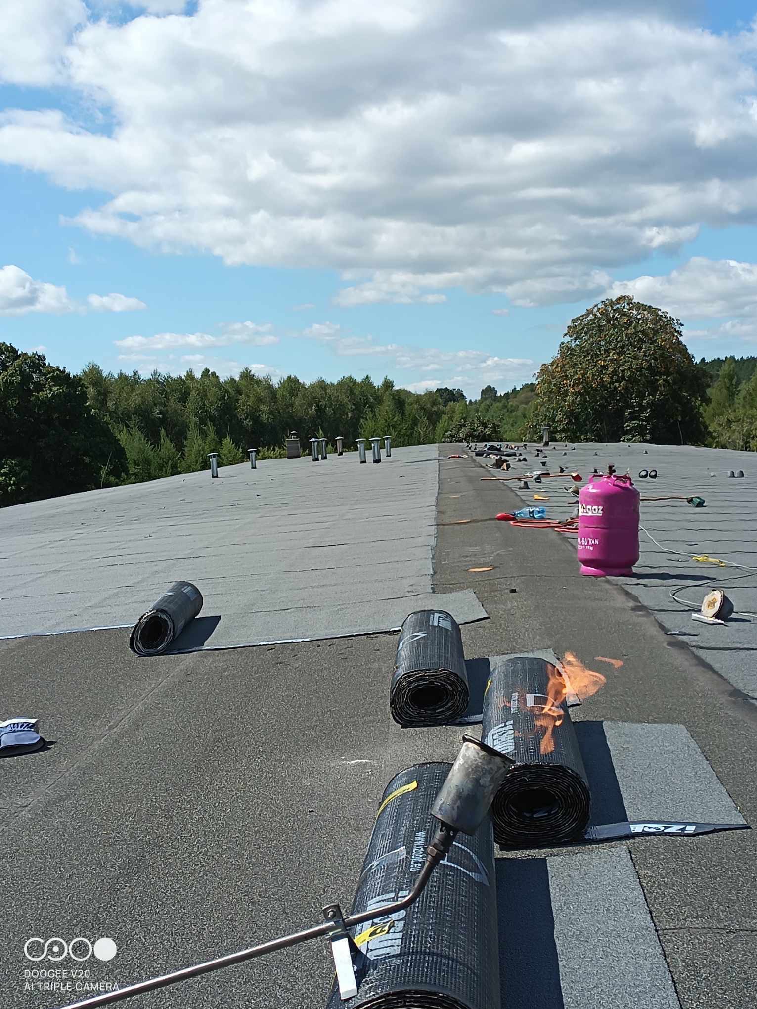 Prace dekarskie na płaskim dachu: palnik dekarski w użyciu, rolki papy, różowa butla gazowa, widok na częściowo pokryty dach i zalesiony horyzont pod błękitnym niebem z chmurami.