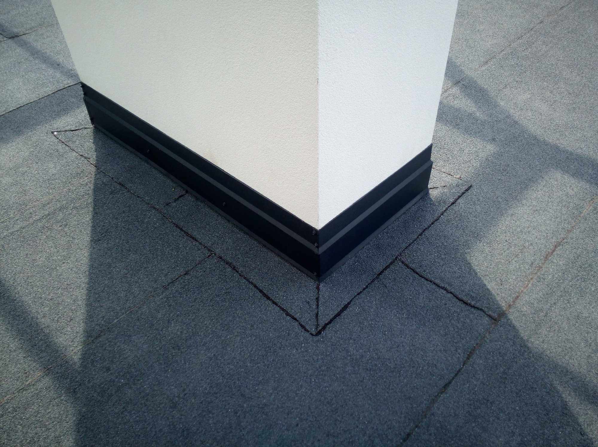 Szczegółowe ujęcie płaskiego dachu pokrytego papą z obróbką blacharską wokół komina, widoczne łączenia papy i czarne listwy wykończeniowe.