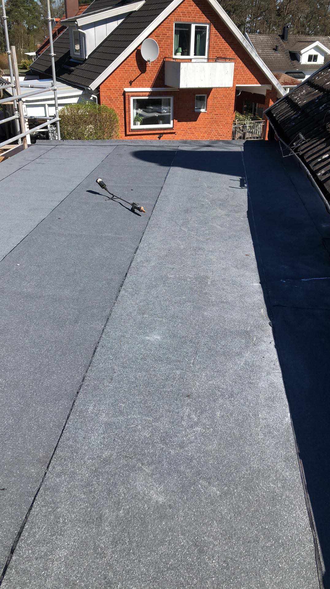 Szary, płaski dach pokryty papą termozgrzewalną, z widocznym palnikiem dekarskim i fragmentem rusztowania w tle.