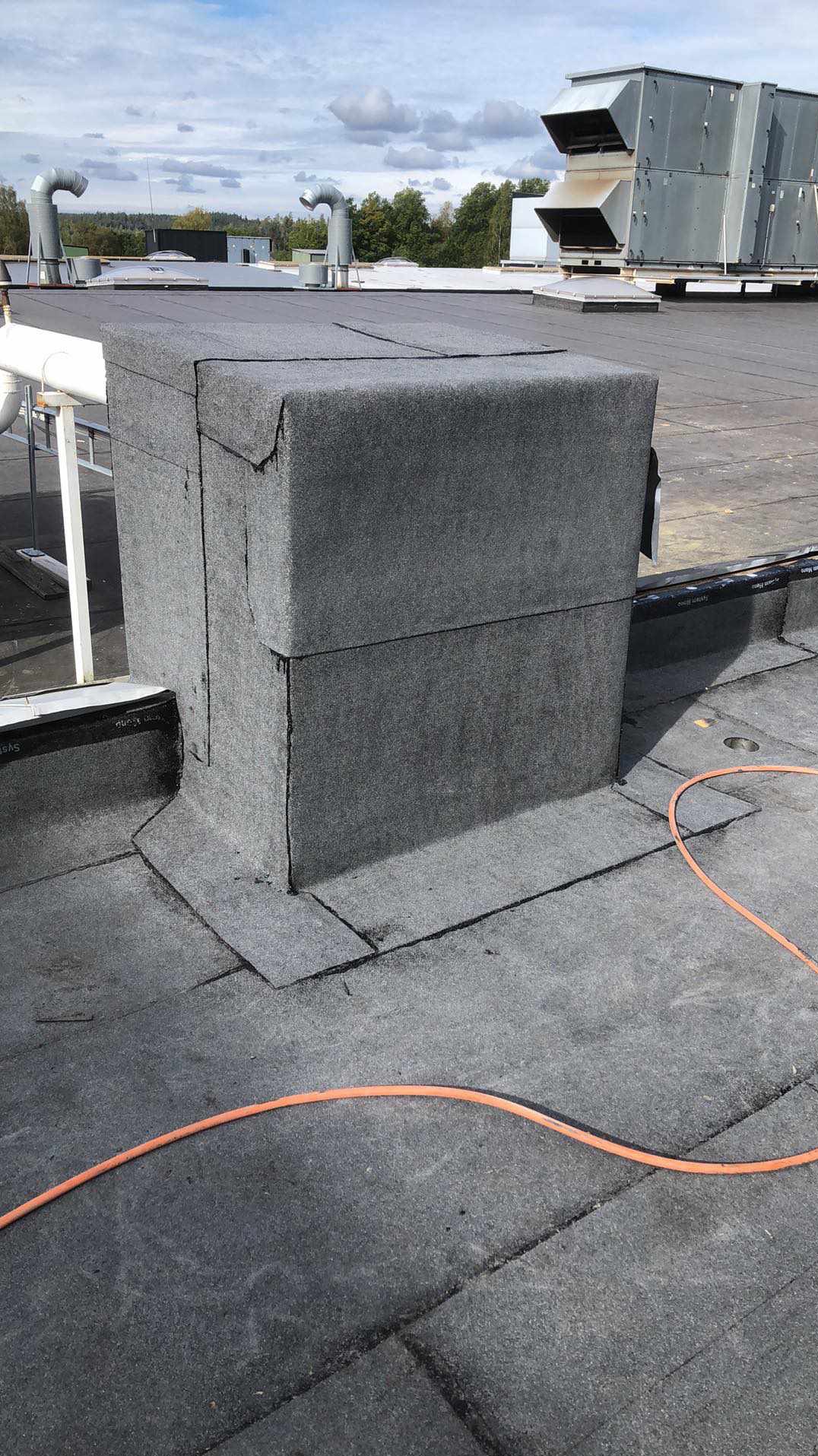 Płaski dach pokryty papą z obróbką komina wentylacyjnego, widoczne elementy wentylacji i pomarańczowy przewód na dachu.