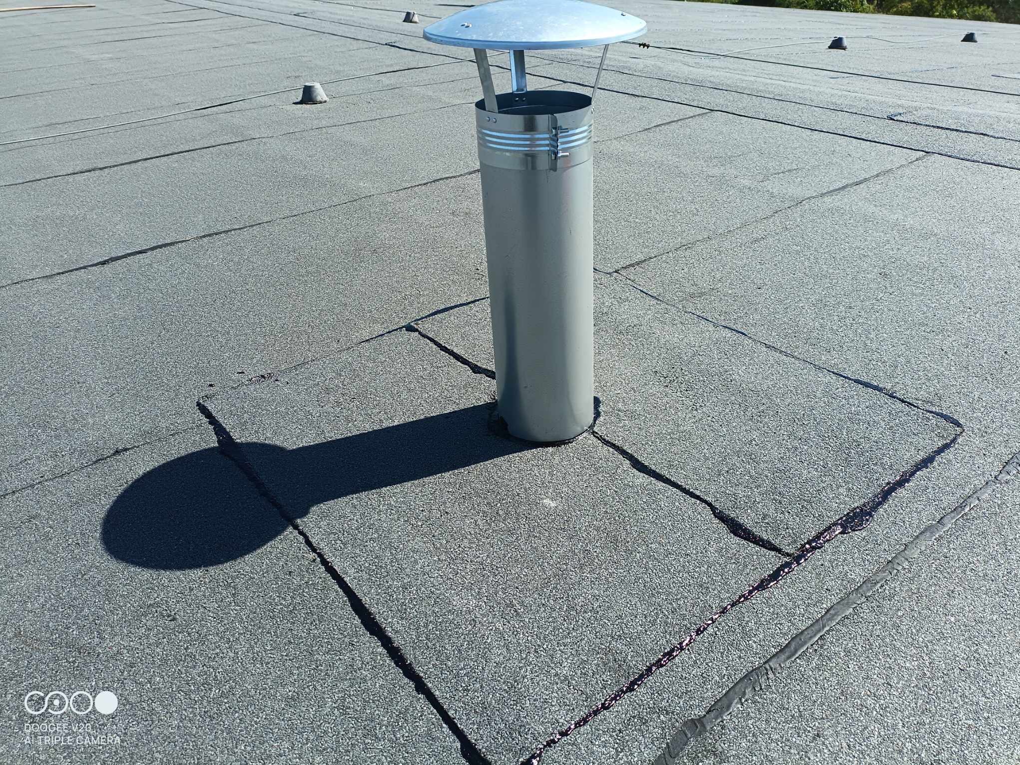 Szary dach pokryty papą z kominem wentylacyjnym; widoczne łączenia papy zalane masą bitumiczną.