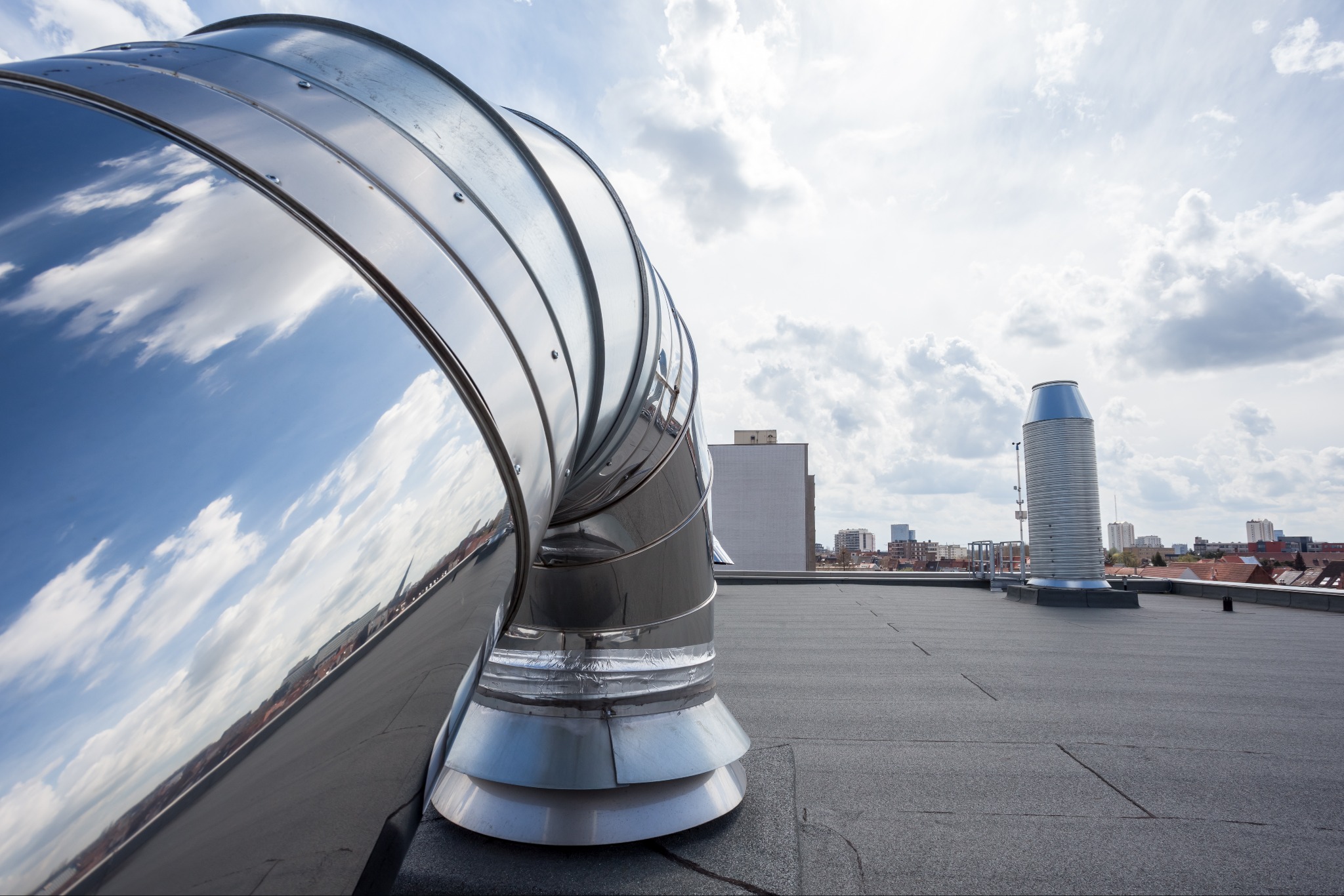 Błyszczący, spiralny element wentylacyjny na płaskim dachu z papy, odbijający błękitne niebo z obłokami, w tle zabudowa miejska.