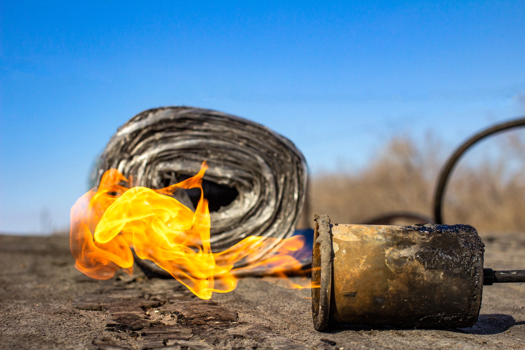 Rozwinięta rolka papy termozgrzewalnej i palnik dekarski z płomieniem na tle błękitnego nieba.
