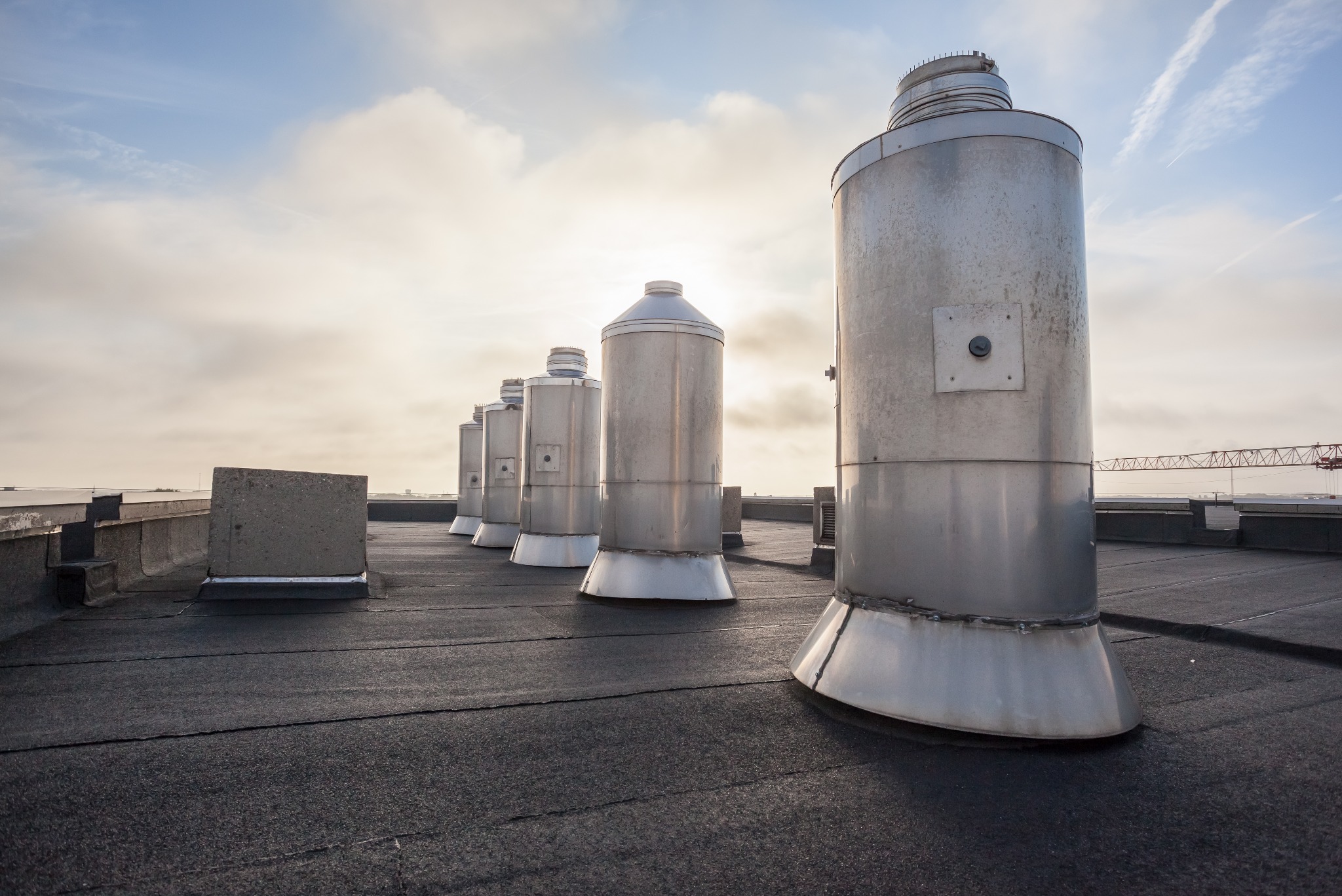 Szereg metalowych kominów wentylacyjnych na płaskim dachu pokrytym papą, z widocznym żurawiem budowlanym w oddali i błękitnym niebem z chmurami.
