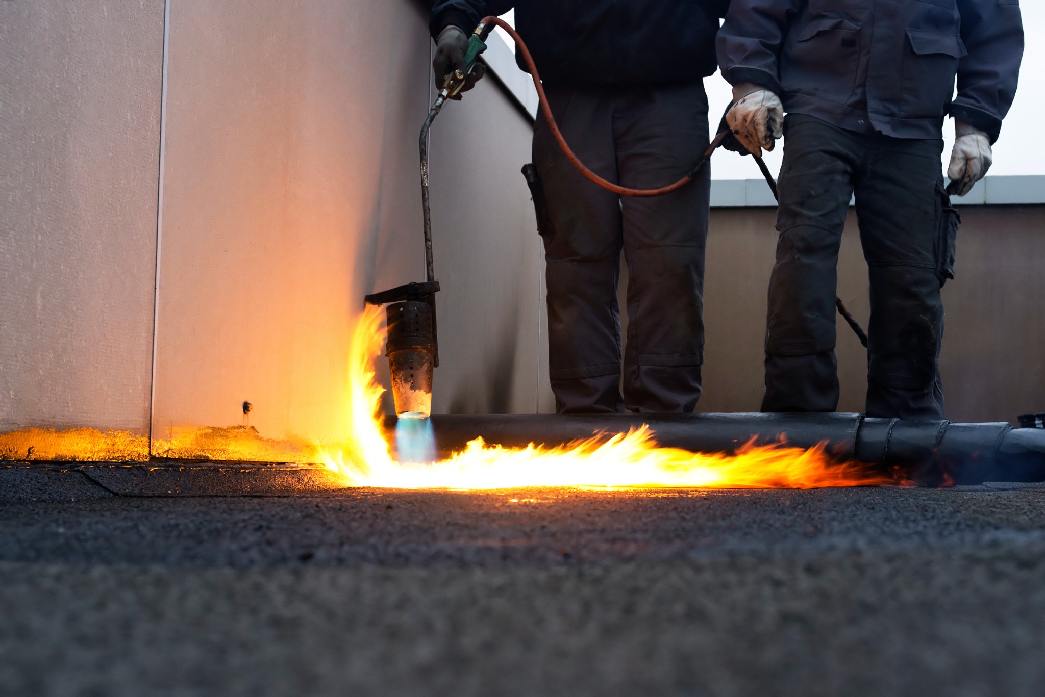 Pracownik w kombinezonie spawa papę na dachu płomieniem palnika na tle ściany budynku.
