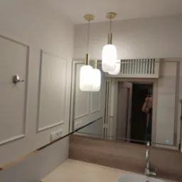 Nowoczesna łazienka z białymi ścianami z dekoracyjnymi ramkami, wiszącymi lampami z elementami w kolorze złota, umywalką wpuszczaną w blat i pralką pod blatem, odbicie osoby robiącej zdjęcie...