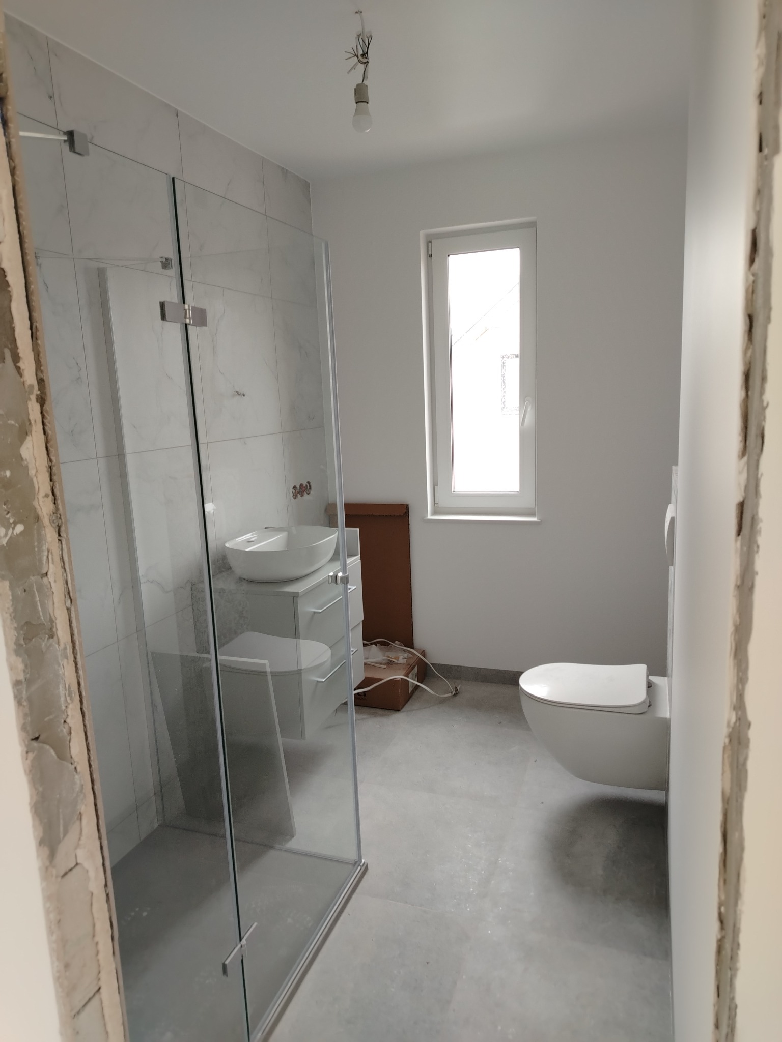 Nowoczesna, jasna łazienka w trakcie wykańczania: kabina prysznicowa ze szklanymi drzwiami, podwieszana toaleta i umywalka nablatowa na szafce, widoczne niedokończone ściany i surowa żarówka.