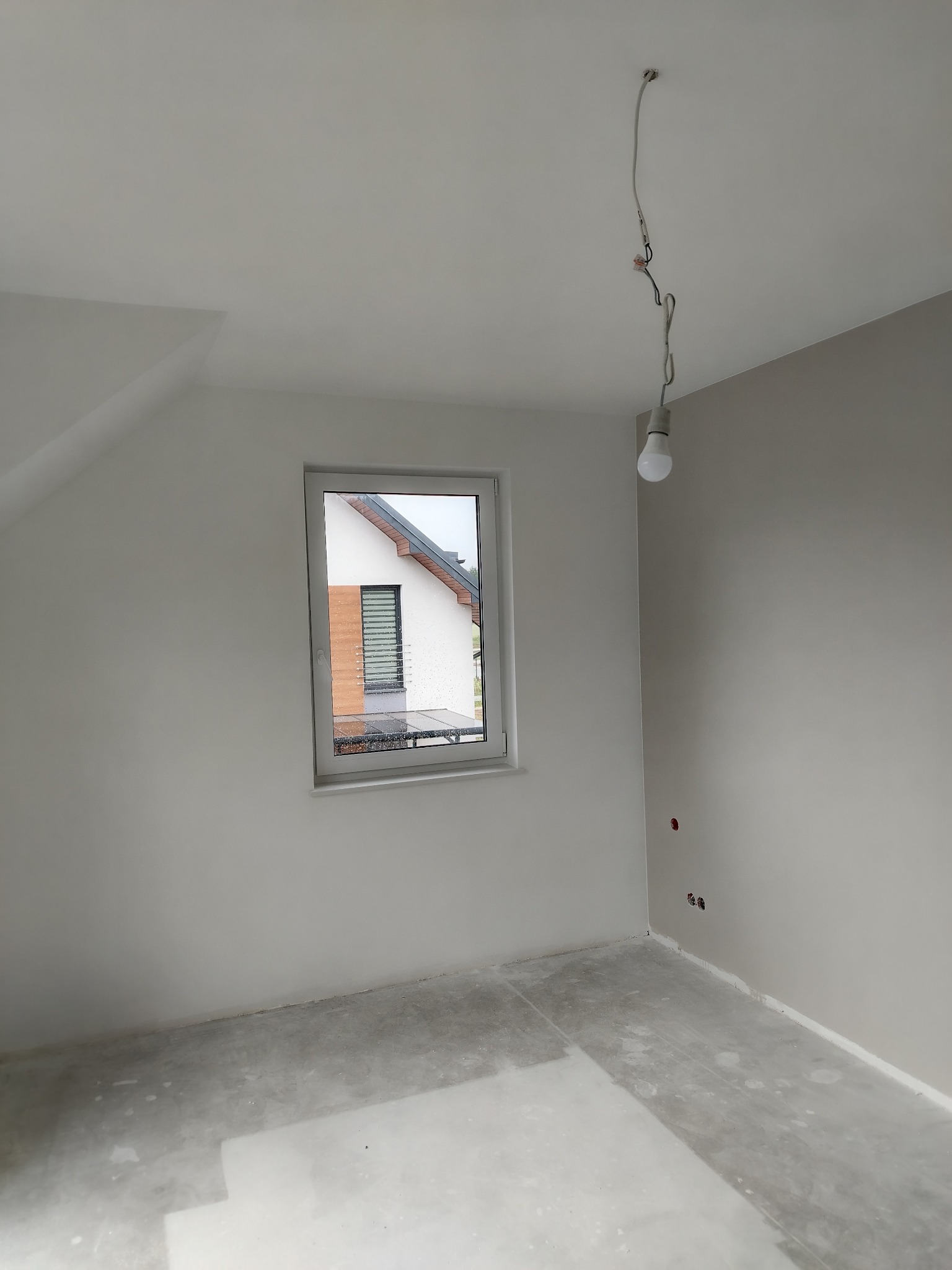 Wnętrze pokoju po szpachlowaniu, z widocznym oknem, wystającymi przewodami żarówki i przygotowanym podłożem pod malowanie.