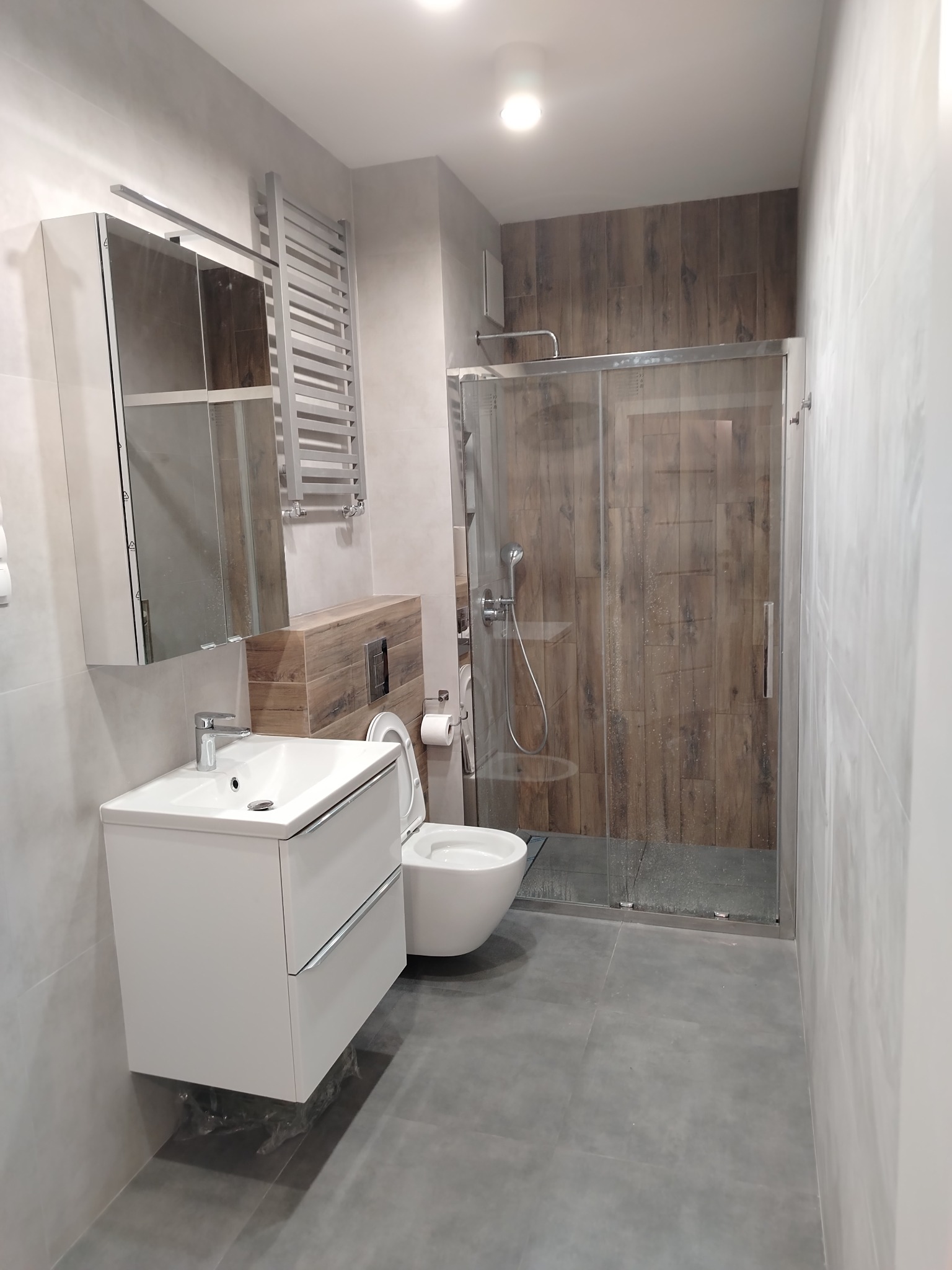 Nowoczesna łazienka z kabiną prysznicową, umywalką z szafką w kolorze białym, grzejnikiem drabinkowym i płytkami imitującymi drewno na ścianie wewnątrz kabiny.