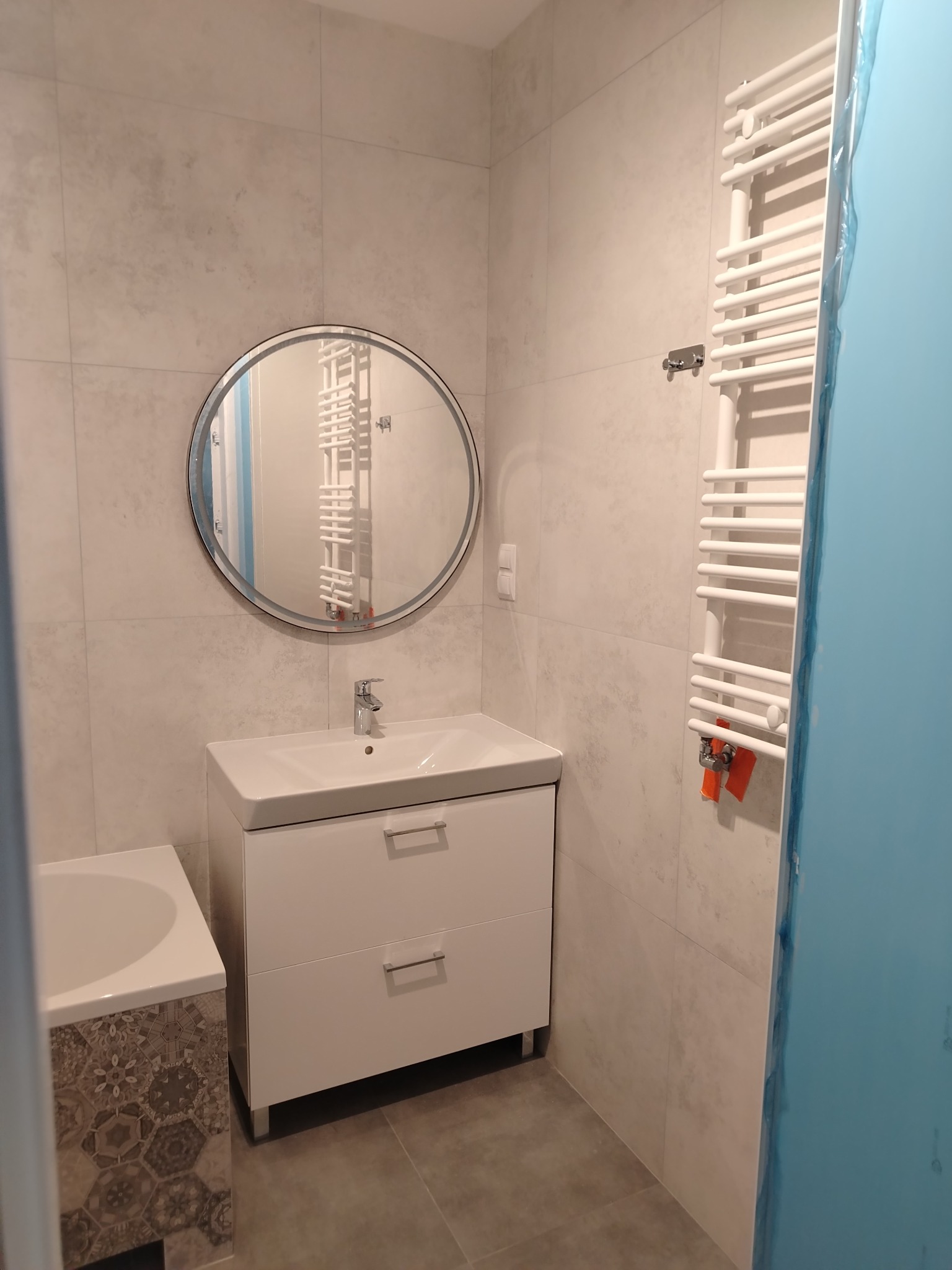 Nowoczesna łazienka z owalnym lustrem nad białą szafką umywalkową, grzejnikiem drabinkowym i płytkami w odcieniach szarości. Fragment wanny z dekoracyjną mozaiką heksagonalną. Zabezpieczenie folią...