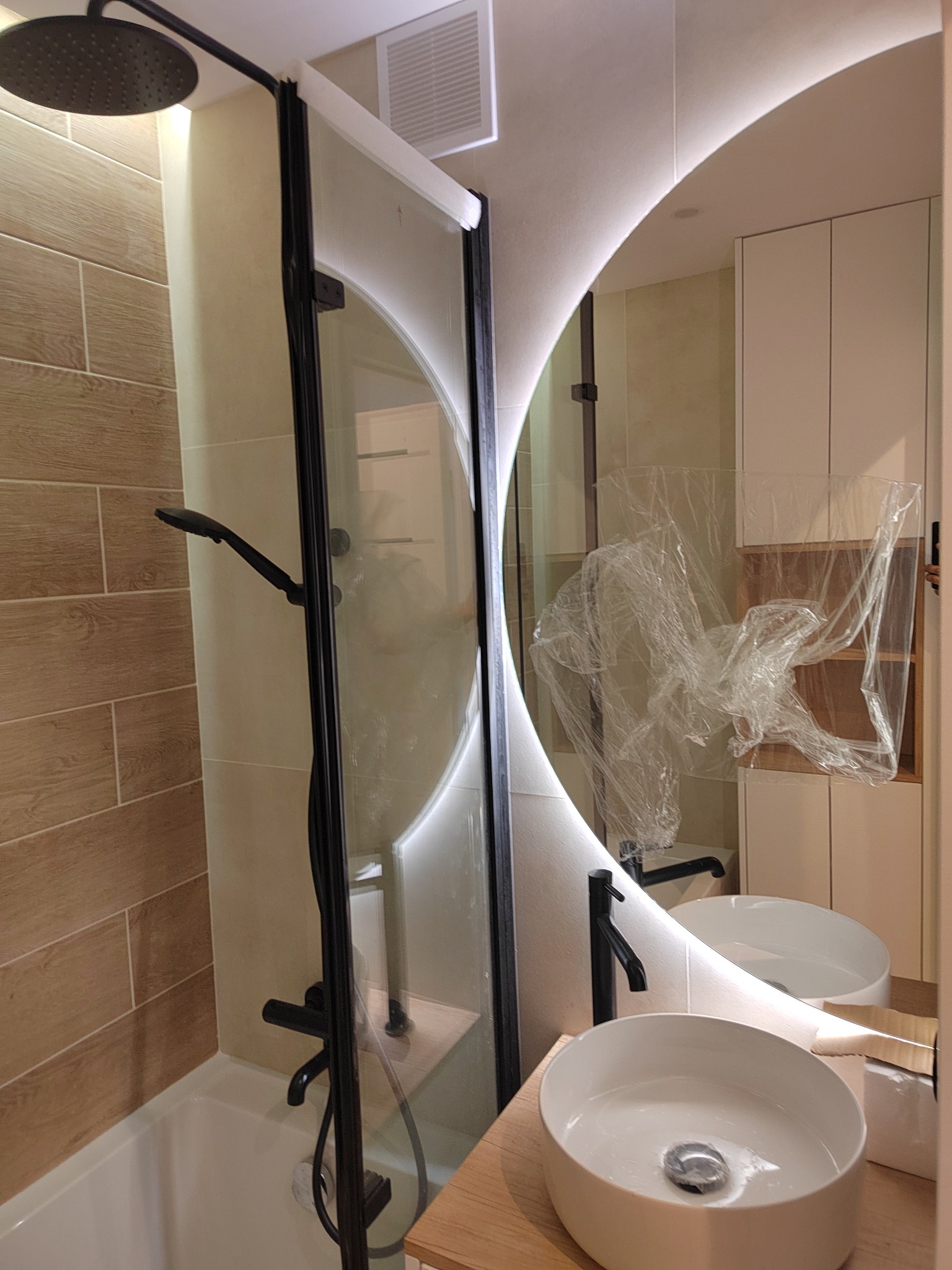 Wykończona łazienka z kabiną prysznicową z czarnymi profilami, okrągłym lustrem z podświetleniem LED i umywalką nablatową. Część szafek w folii ochronnej.