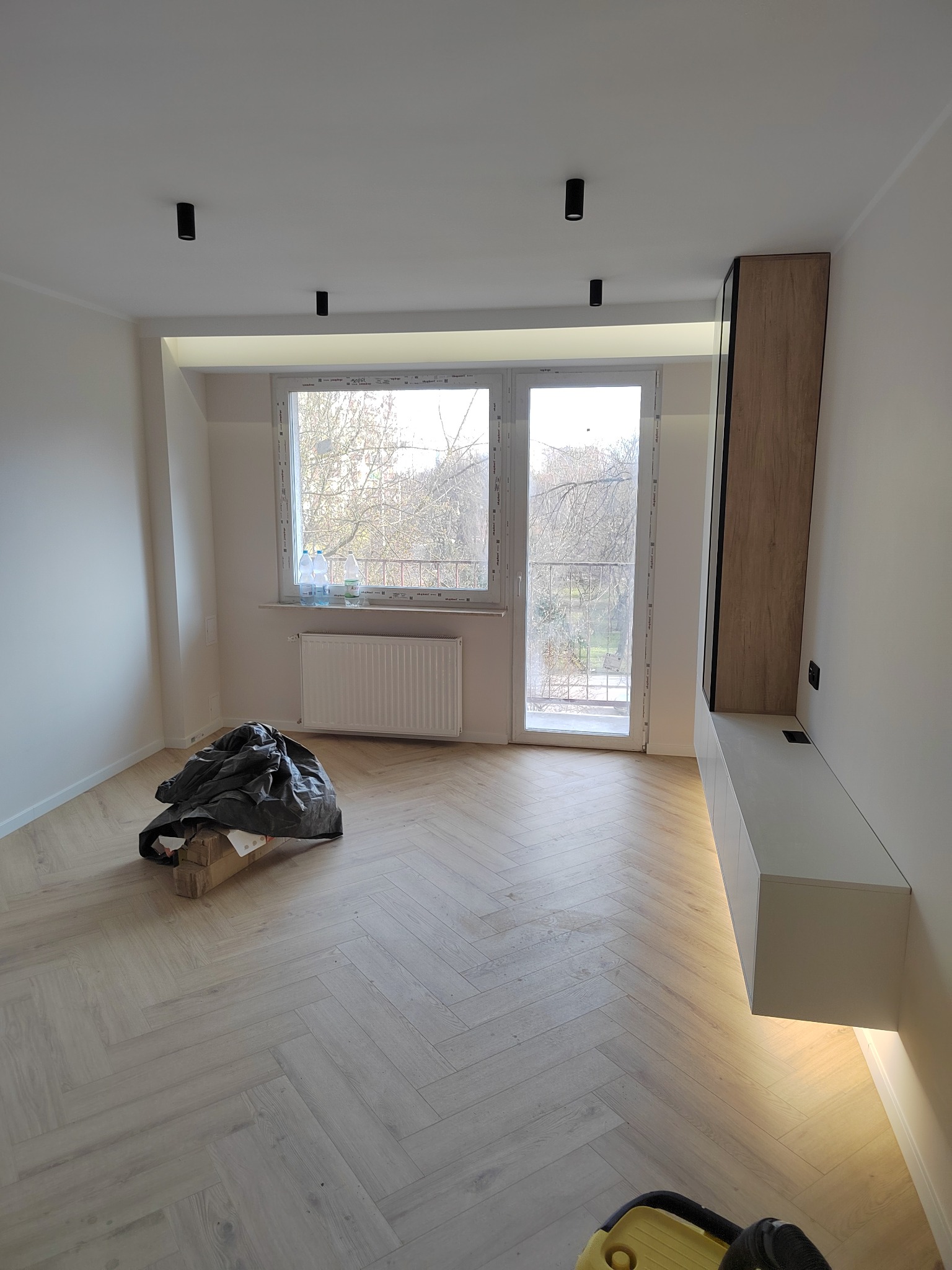Jasny pokój po remoncie z parkietem ułożonym w jodełkę, z workiem gruzu pod oknem i zabudową meblową z podświetleniem LED.
