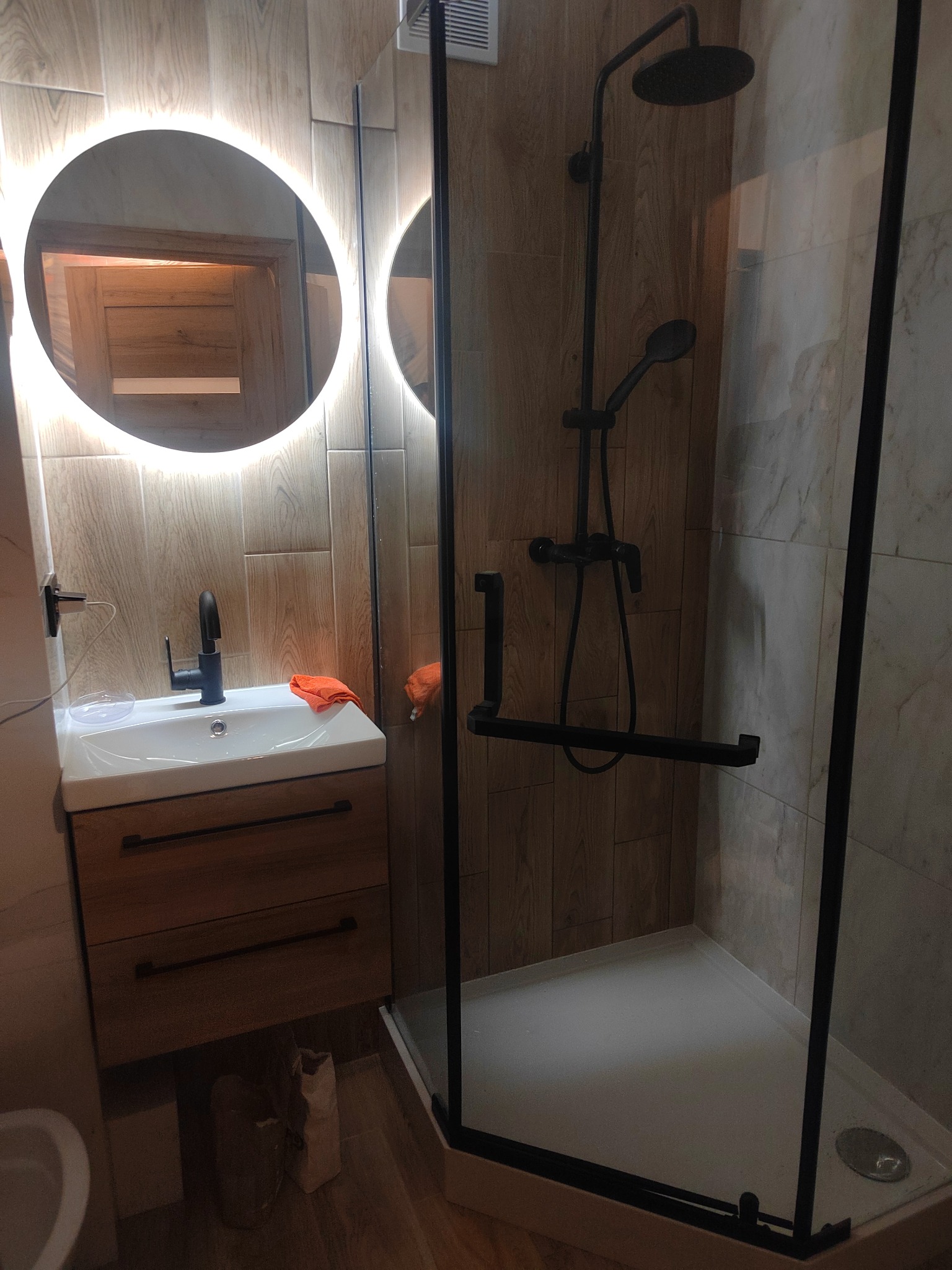 Nowoczesna łazienka z kabiną prysznicową typu walk-in z czarną armaturą, okrągłym lustrem z podświetleniem LED i drewnopodobnymi płytkami na ścianach.