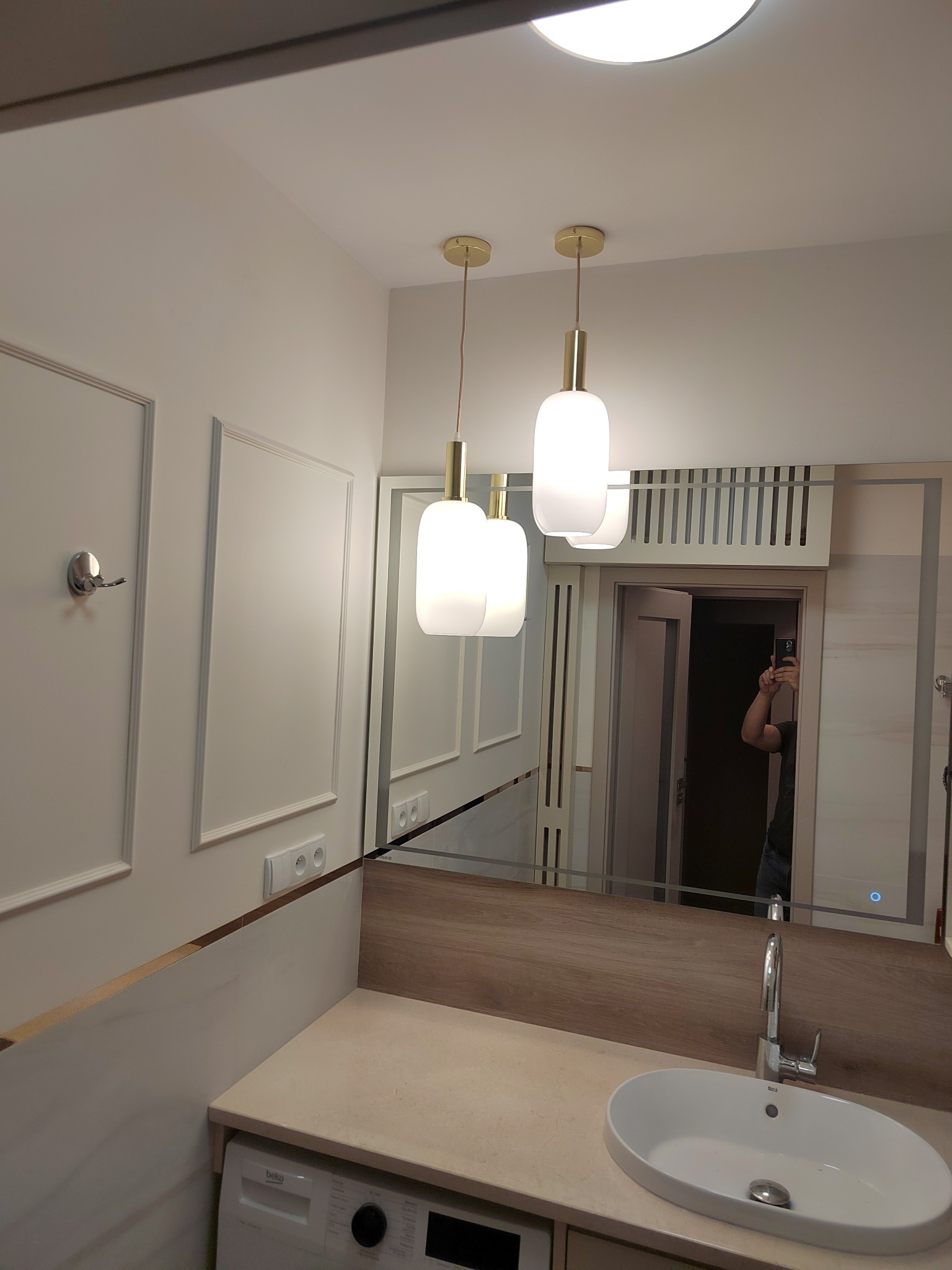 Nowoczesna łazienka z białymi ścianami z dekoracyjnymi ramkami, wiszącymi lampami z elementami w kolorze złota, umywalką wpuszczaną w blat i pralką pod blatem, odbicie osoby robiącej zdjęcie...