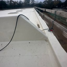 PRO-BUD DANIEL BOŁOTOW - Biała membrana dachowa z widocznymi świetlikami, przykrywająca płaski dach, z kablem zasilającym i złączką elektryczną na krawędzi, na tle pochmurnego nieba i drzew.