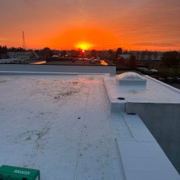 PRO-BUD DANIEL BOŁOTOW - Biały dach płaski z świetlikiem i zieloną skrzynką narzędziową Leister na tle pomarańczowego wschodu słońca nad miastem.