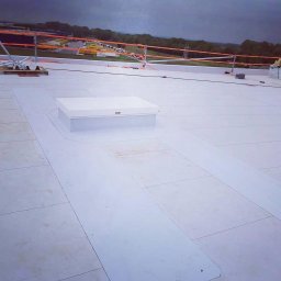 PRO-BUD DANIEL BOŁOTOW - Białe panele izolacyjne na płaskim dachu z świetlikiem dachowym, widoczne zabezpieczenia pomarańczową barierką i oddalona zabudowa.