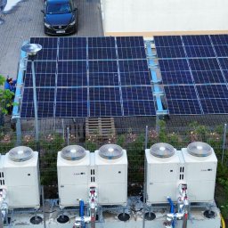 PRO-BUD DANIEL BOŁOTOW - Instalacja fotowoltaiczna i pompy ciepła na zewnątrz budynku, widok z góry. Na pierwszym planie 4 pompy ciepła, w tle panele fotowoltaiczne i pracownik w niebieskiej kurtce.