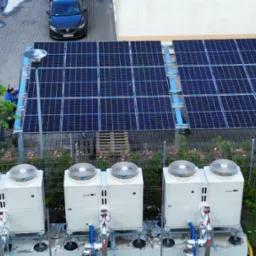 Instalacja fotowoltaiczna i pompy ciepła na zewnątrz budynku, widok z g&oacute;ry. Na pierwszym planie 4 pompy ciepła, w tle panele fotowoltaiczne i pracownik w niebieskiej kurtce.