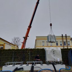 PRO-BUD DANIEL BOŁOTOW - Montaż pompy ciepła z użyciem dźwigu. Widoczne fundamenty, rury i pracownik nadzorujący. Urządzenie jeszcze w folii ochronnej, przygotowane do instalacji.