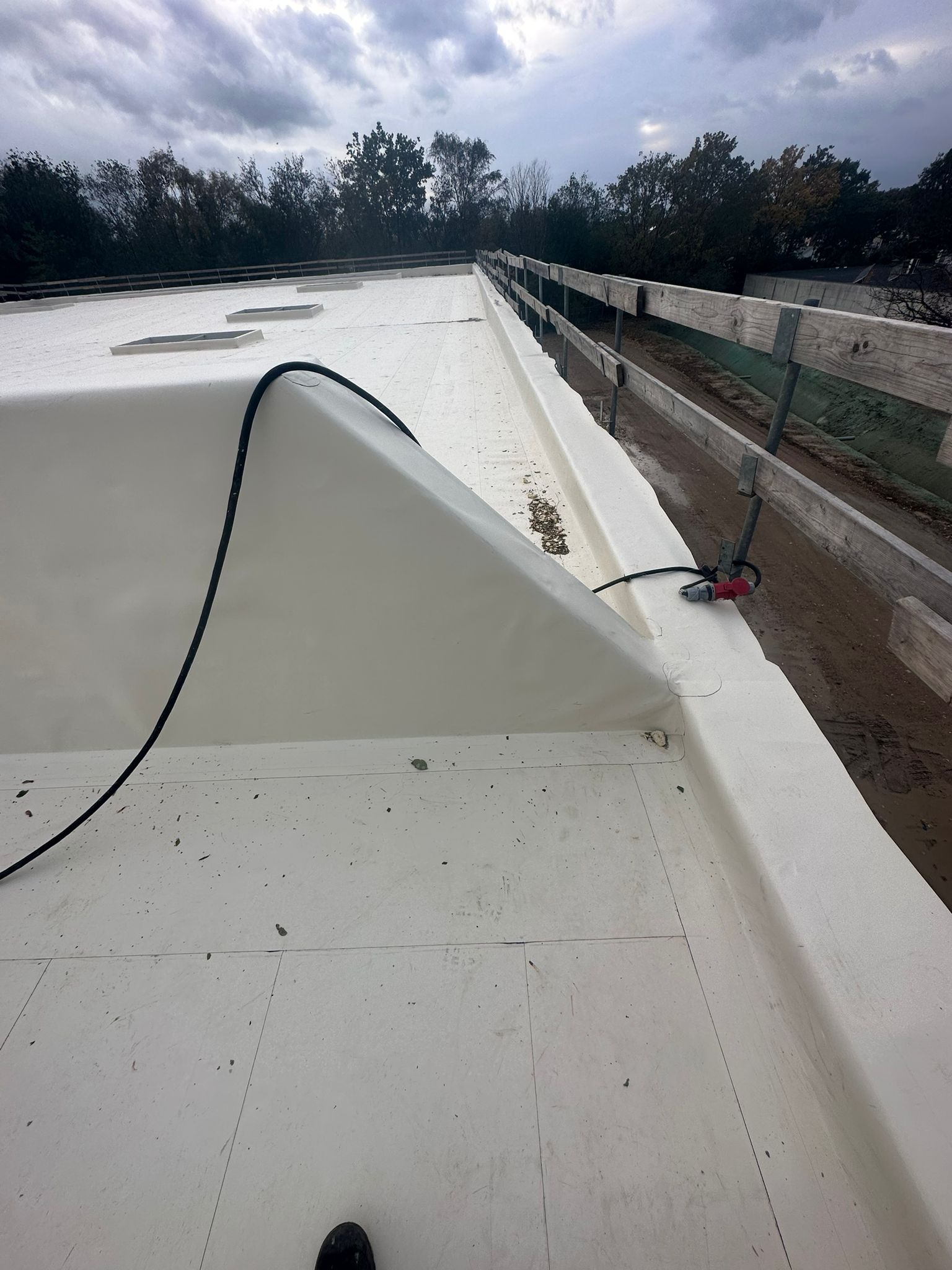 Biała membrana dachowa z widocznymi świetlikami, przykrywająca płaski dach, z kablem zasilającym i złączką elektryczną na krawędzi, na tle pochmurnego nieba i drzew.