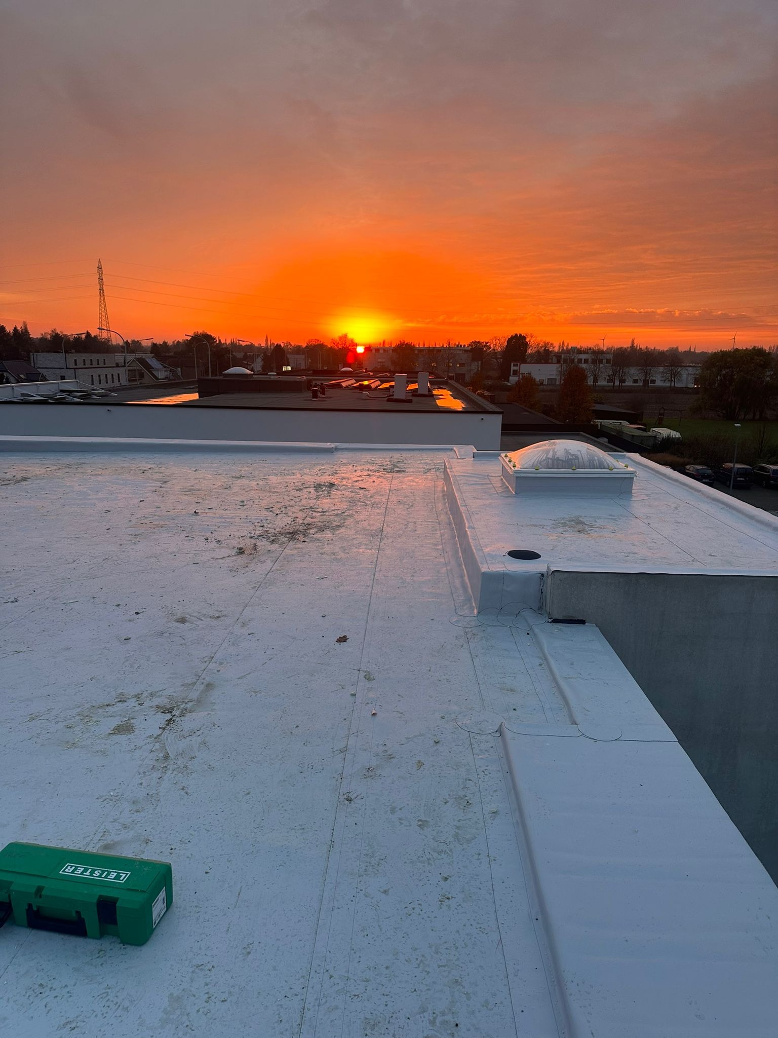 Biały dach płaski z świetlikiem i zieloną skrzynką narzędziową Leister na tle pomarańczowego wschodu słońca nad miastem.