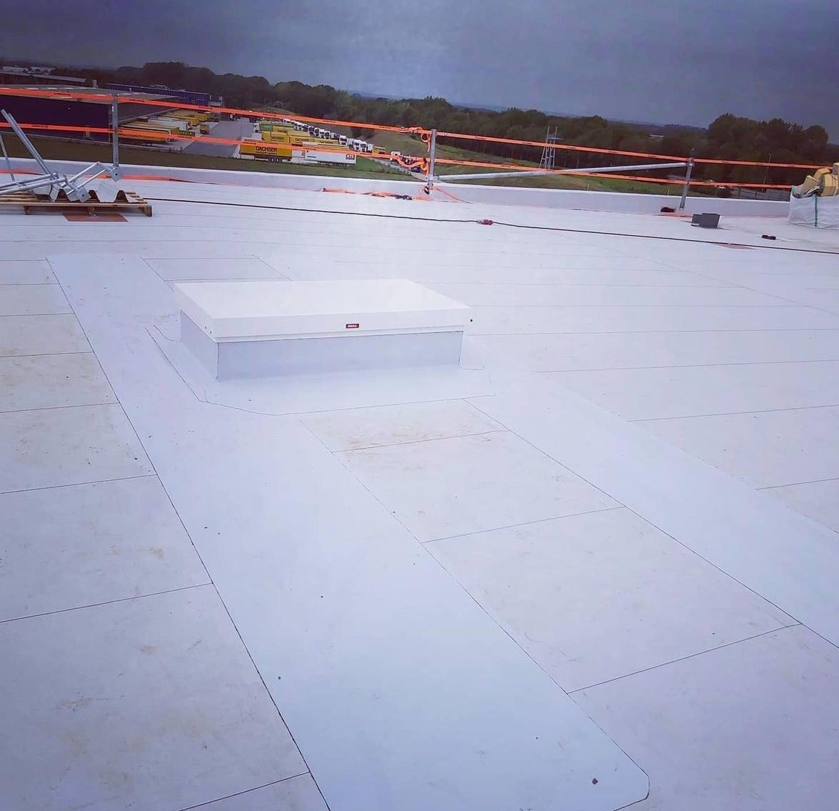 Białe panele izolacyjne na płaskim dachu z świetlikiem dachowym, widoczne zabezpieczenia pomarańczową barierką i oddalona zabudowa.