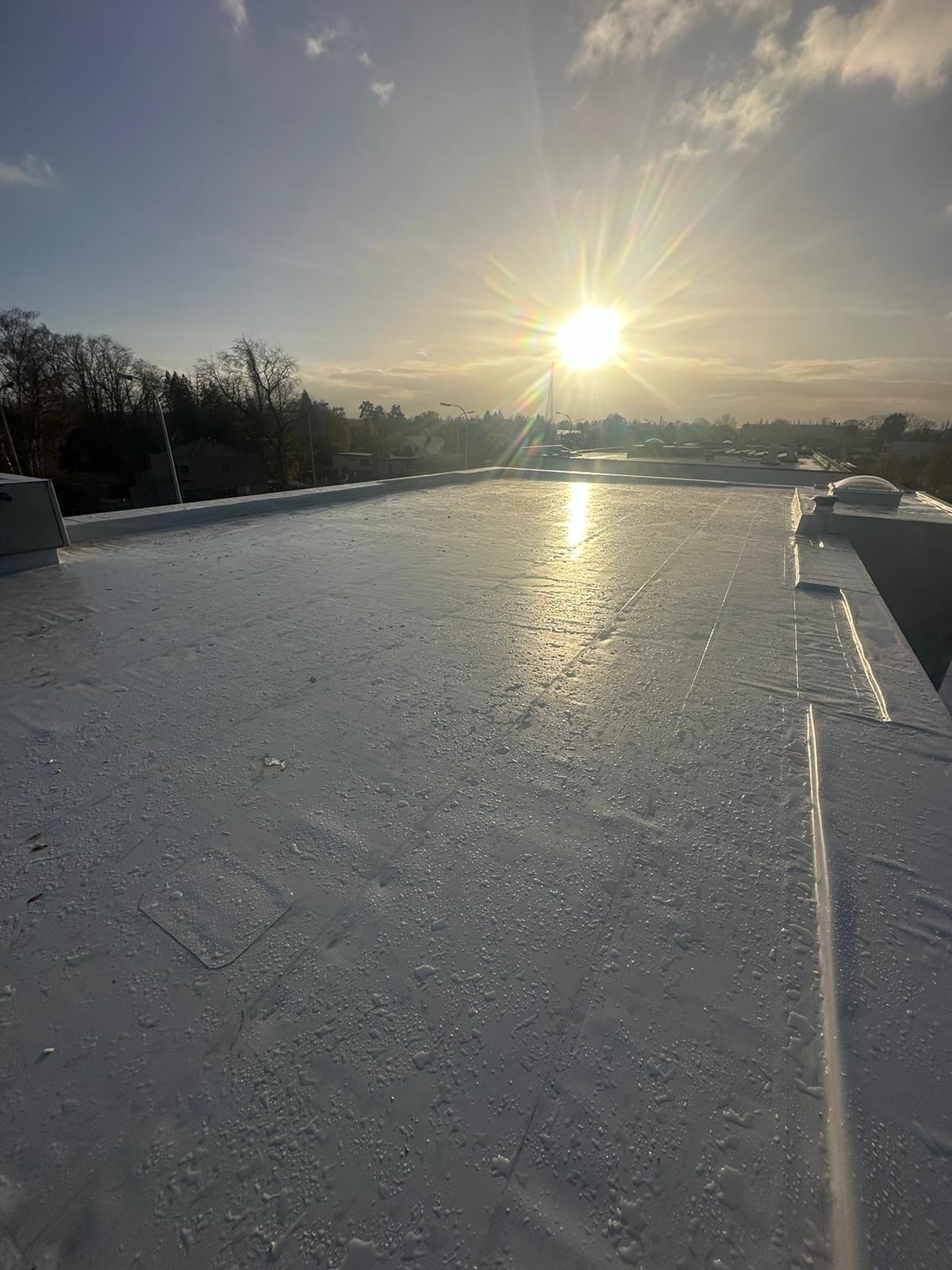 Biała, płaska powierzchnia dachu z widocznymi śladami wilgoci, oświetlona promieniami wschodzącego słońca, w tle drzewa i zabudowania.