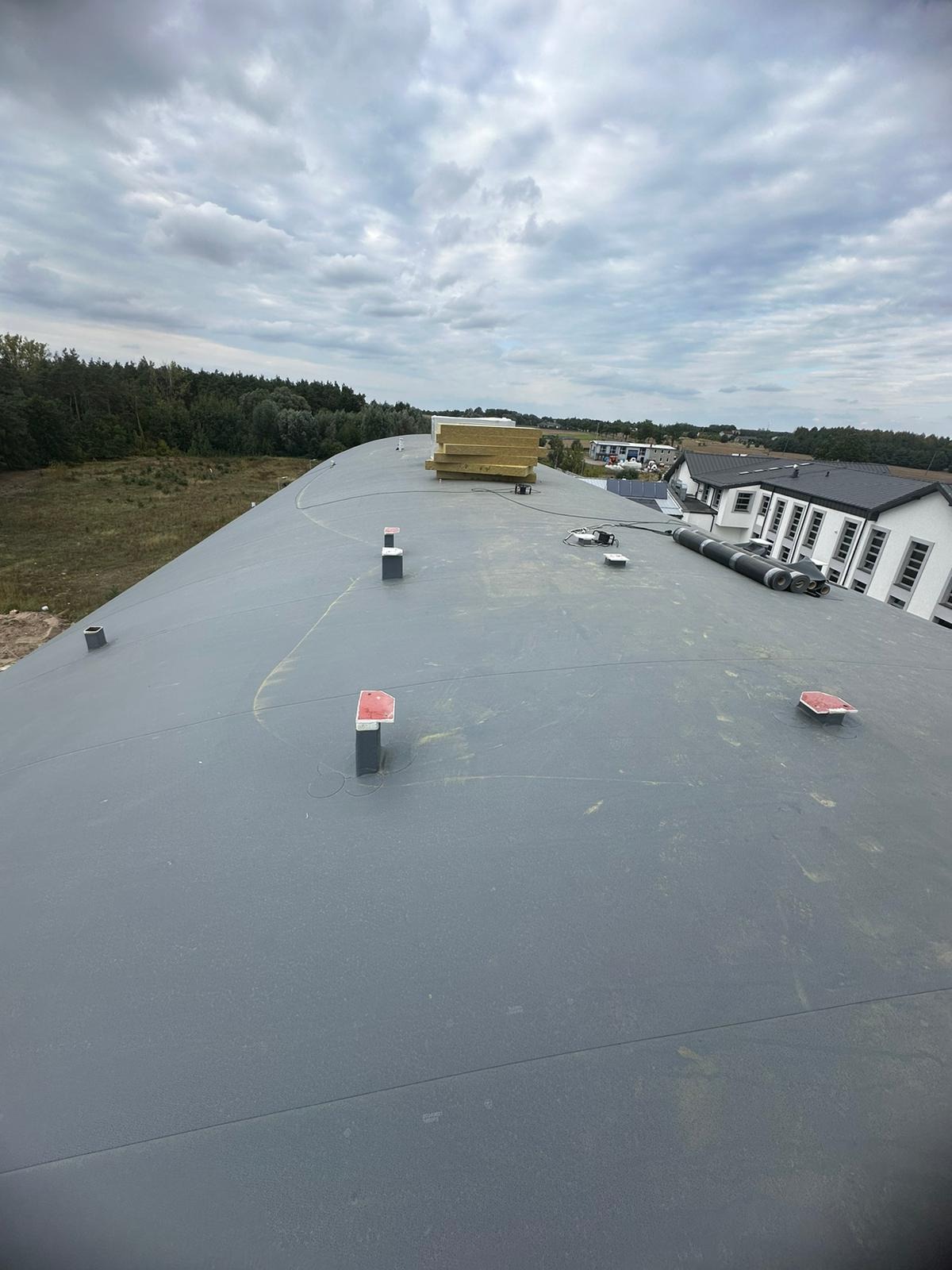 Szary, płaski dach z widocznymi kominami wentylacyjnymi z czerwonymi daszkami, ułożone płyty izolacyjne oraz rury wentylacyjne, w tle zabudowa jednorodzinna i drzewa