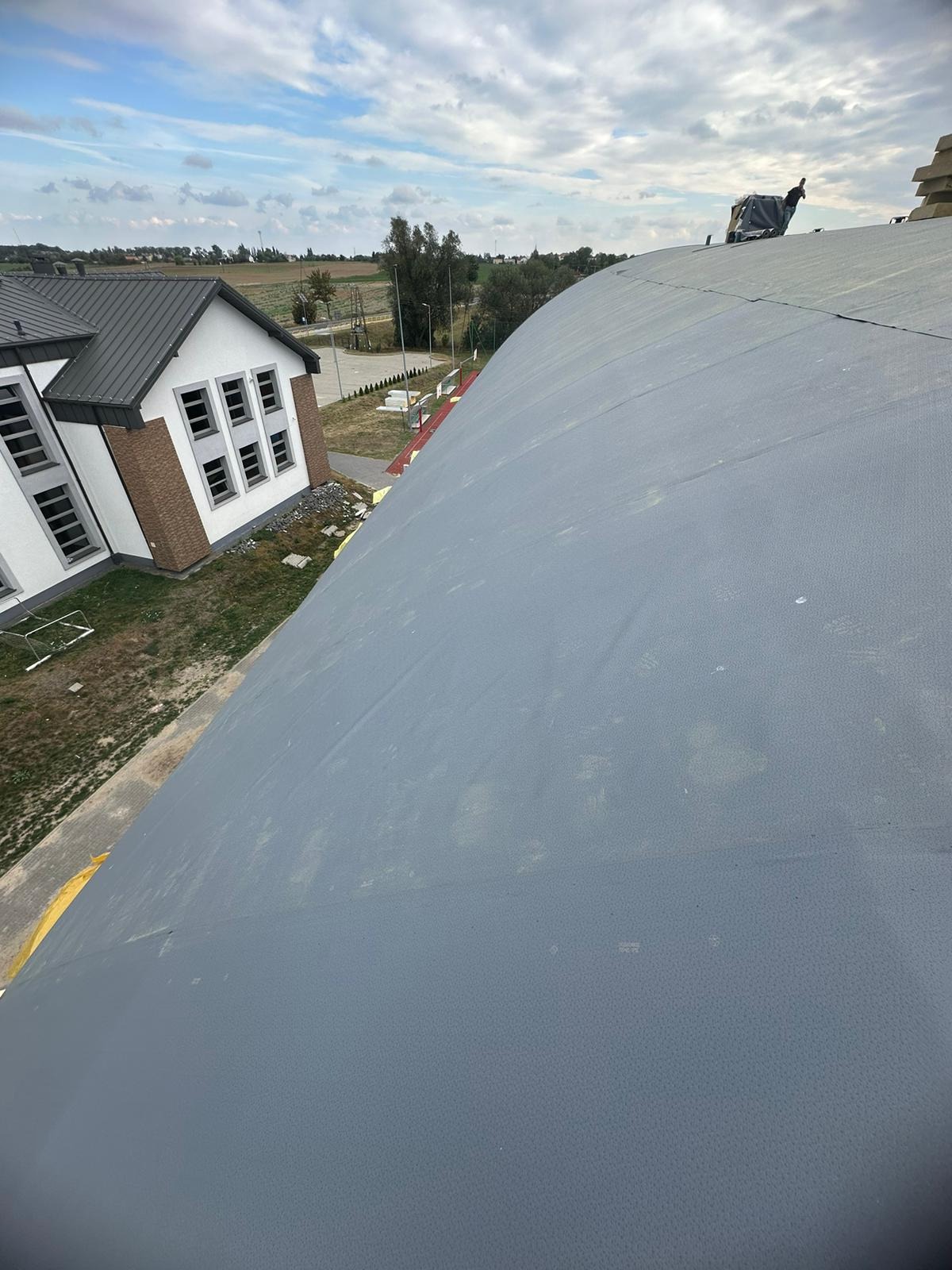 Szare, zaokrąglone pokrycie dachu hali sportowej z widocznym fragmentem elewacji budynku obok i pracownikiem na dachu w oddali, w tle pola i niebo z chmurami.