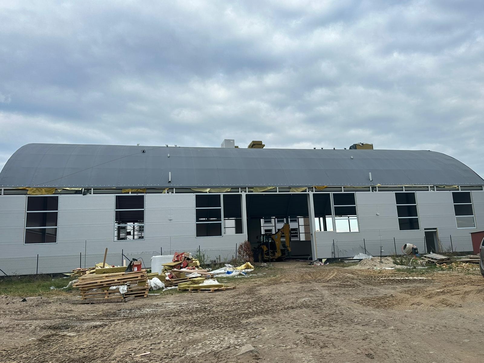 Budynek przemysłowy w trakcie budowy lub ocieplania z widocznym sprzętem budowlanym i materiałami izolacyjnymi na zewnątrz, pochmurne niebo w tle.