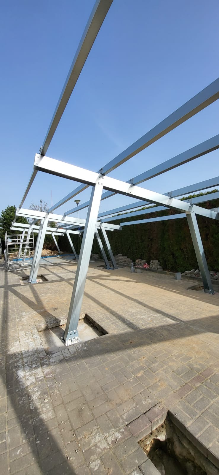 Stalowa konstrukcja wsporcza na kostce brukowej, prawdopodobnie pod panele fotowoltaiczne, widoczne otwory w ziemi pod fundamenty.