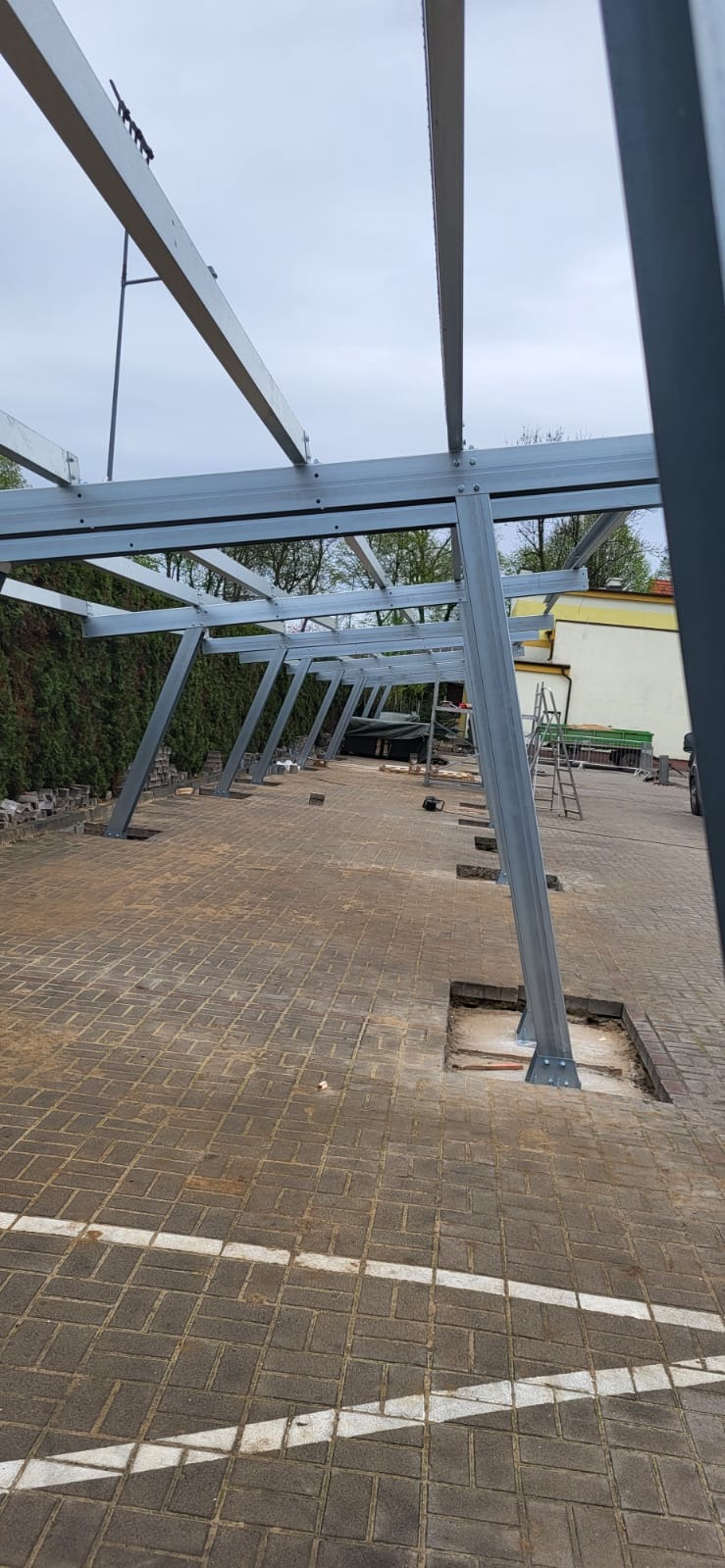 Montaż stalowej konstrukcji pod panele fotowoltaiczne na posesji. Widoczne fundamenty i częściowo zamontowane elementy szkieletu. Kostka brukowa z wyciętymi otworami.