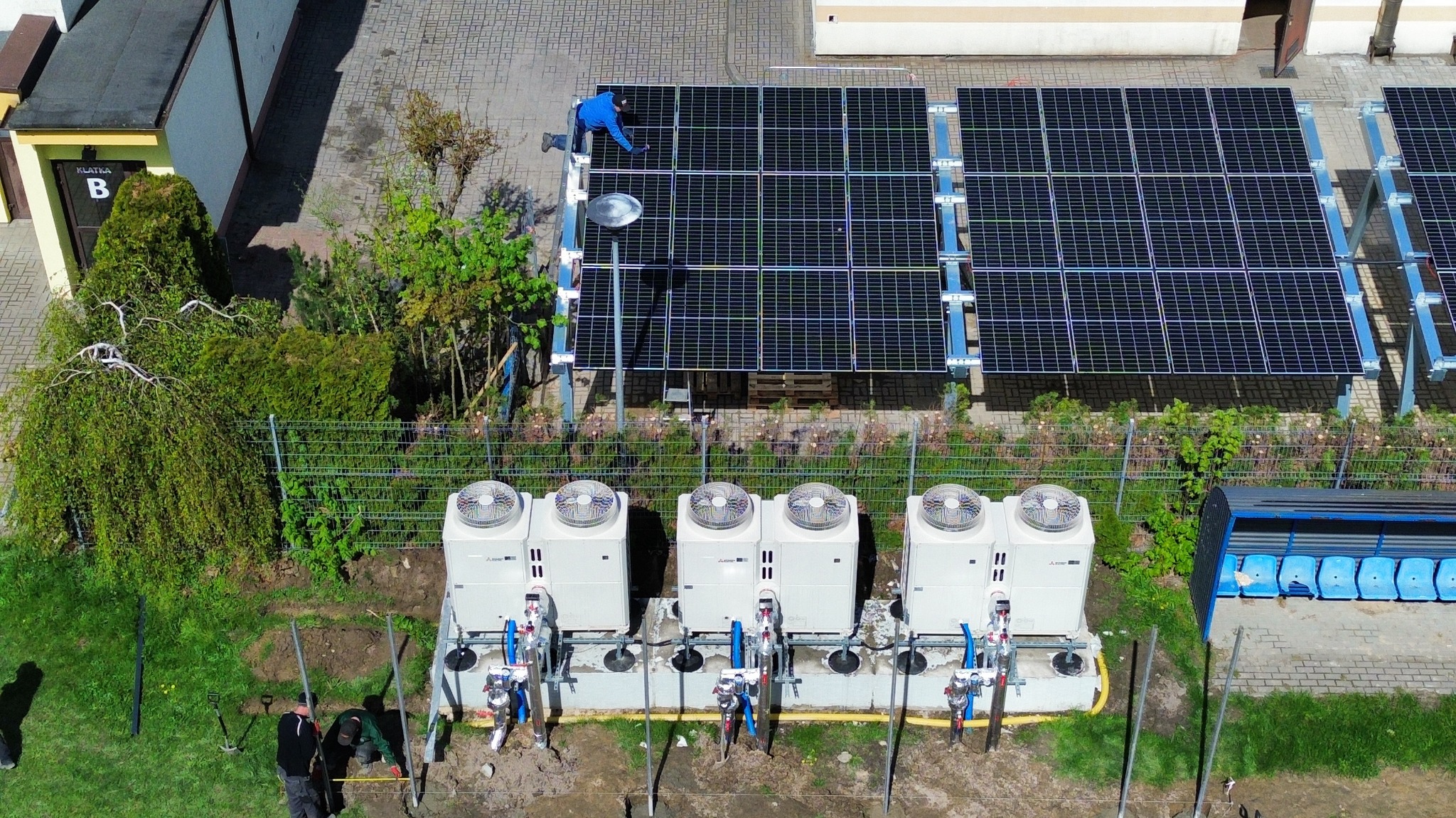 Montaż instalacji z panelami słonecznymi i pompami ciepła. Widok z góry ukazuje prace instalacyjne, panele oraz urządzenia w otoczeniu zieleni.