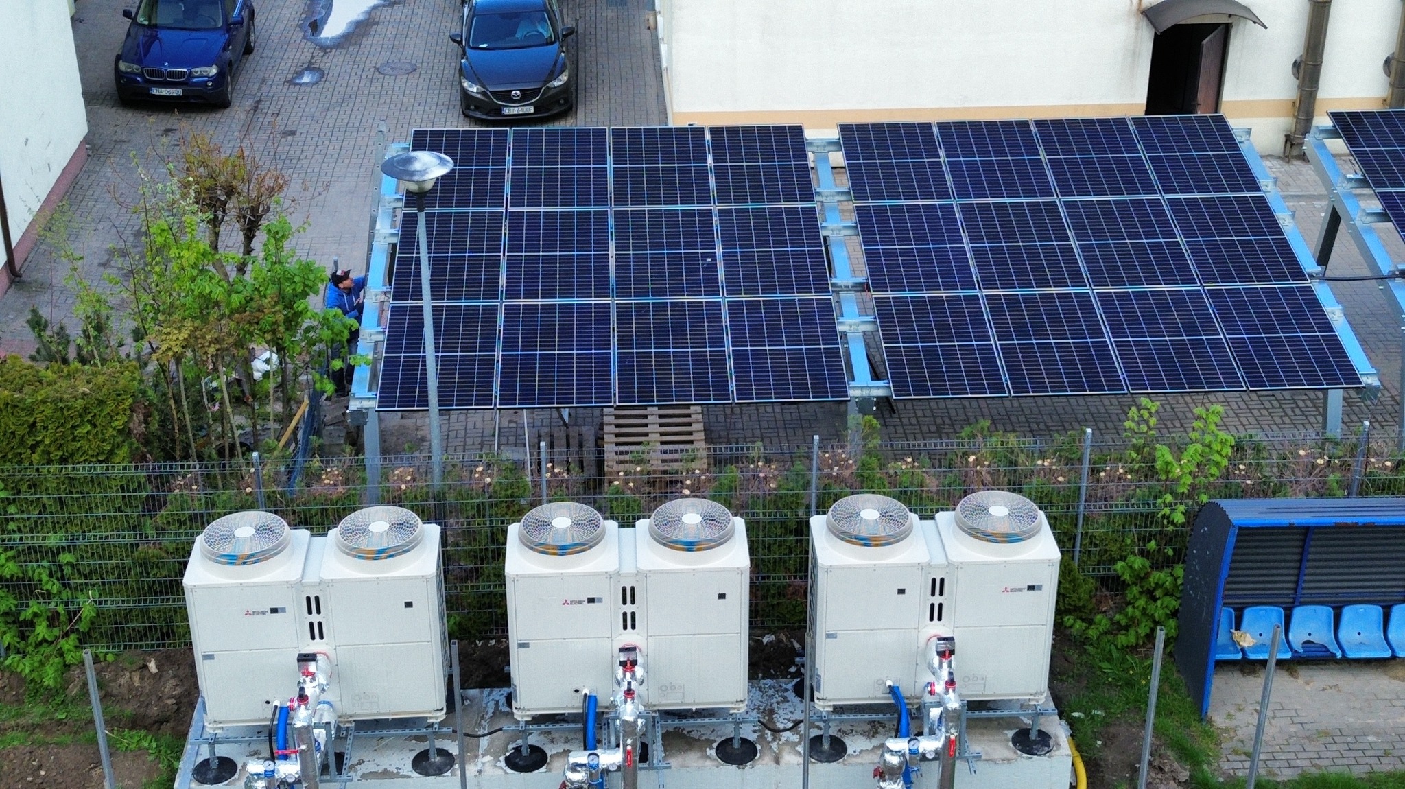 Instalacja fotowoltaiczna i pompy ciepła na zewnątrz budynku, widok z góry. Na pierwszym planie 4 pompy ciepła, w tle panele fotowoltaiczne i pracownik w niebieskiej kurtce.