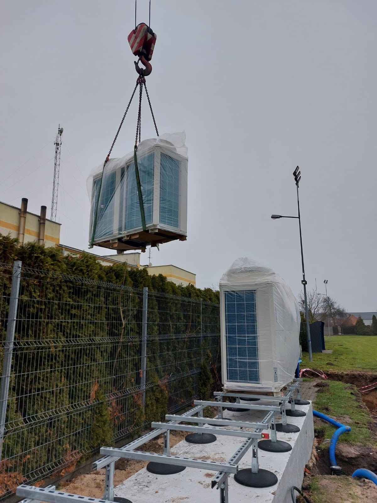 Montaż pompy ciepła na przygotowanym fundamencie za pomocą dźwigu. Urządzenie w folii ochronnej. Widoczny fragment ogrodzenia i budynek w tle.