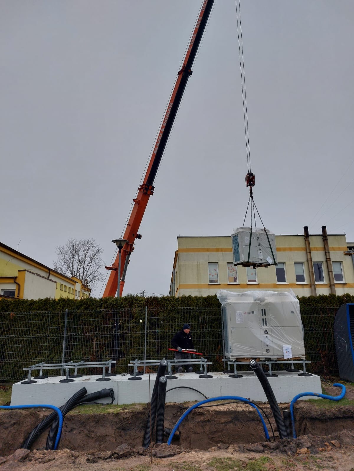 Montaż pompy ciepła z użyciem dźwigu. Widoczne fundamenty, rury i pracownik nadzorujący. Urządzenie jeszcze w folii ochronnej, przygotowane do instalacji.