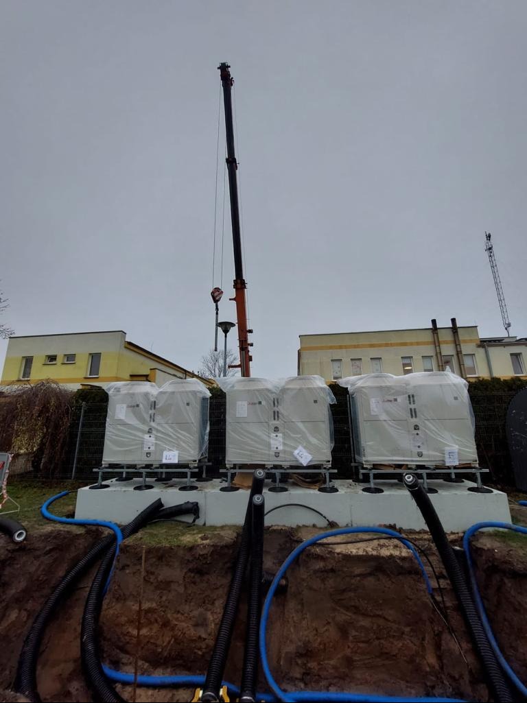Montaż trzech pomp ciepła na betonowym fundamencie. Widoczny dźwig, rury w wykopie i budynki w tle. Pompy częściowo owinięte folią.