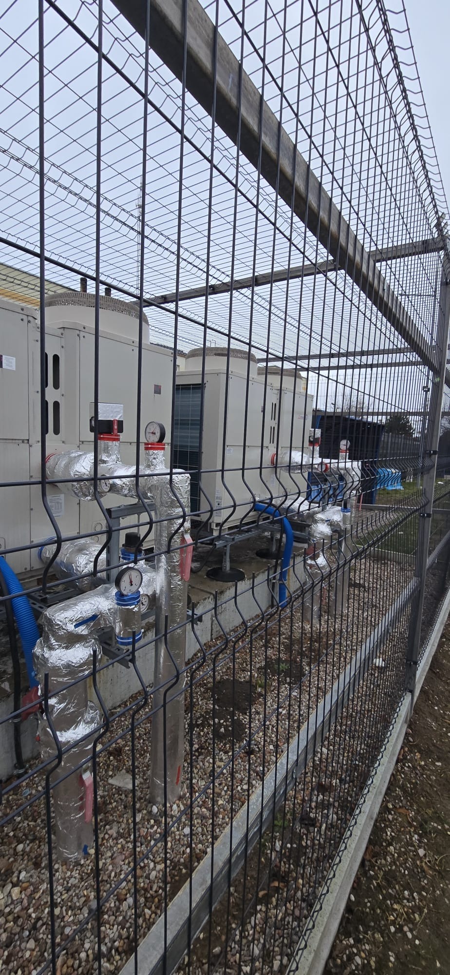 Szereg jednostek zewnętrznych pompy ciepła za ogrodzeniem z siatki, widoczne rury w izolacji aluminiowej i manometry. Szary dzień, instalacja na zewnątrz budynku.