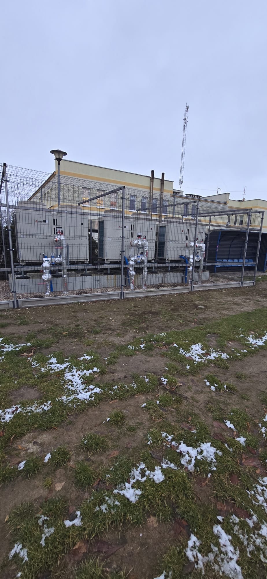 Zewnętrzna instalacja kilku pomp ciepła Mitsubishi Electric z rurami w izolacji, zabezpieczona ogrodzeniem z siatki na tle budynku w pochmurny dzień.