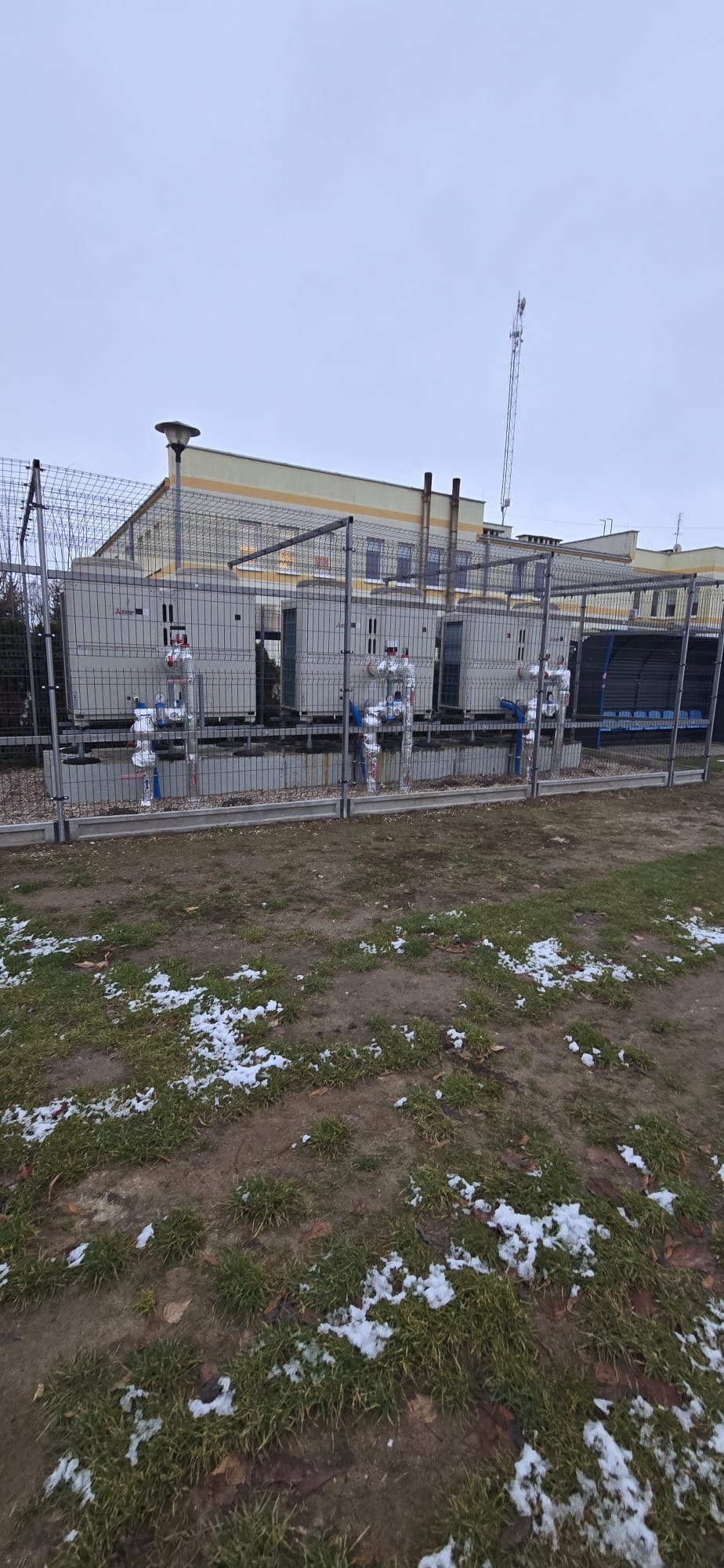 Instalacja kilku zewnętrznych jednostek pompy ciepła Mitsubishi, zabezpieczonych ogrodzeniem z siatki na tle budynku. Widoczne rury i zawory.