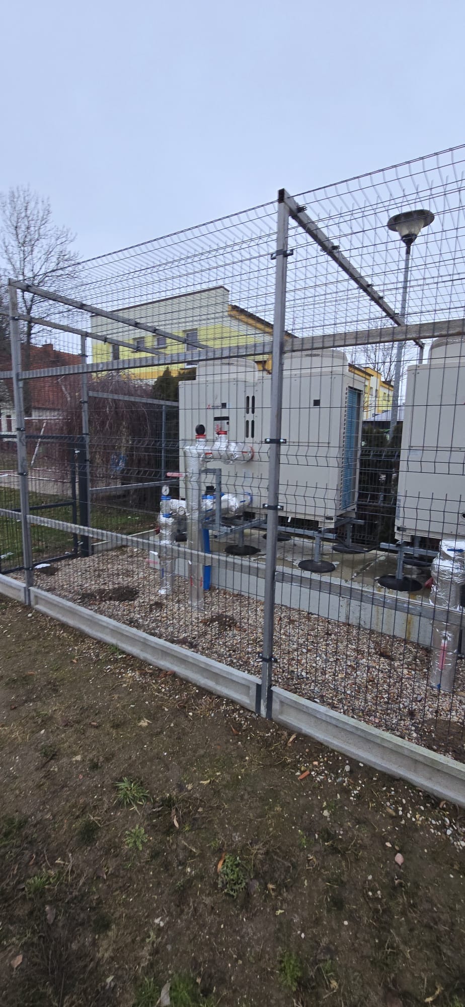 Zabezpieczona siatką instalacja dwóch białych pomp ciepła na zewnątrz budynku. Widoczne rury, zawory, manometry i elementy montażowe. Tło to budynki i drzewa.