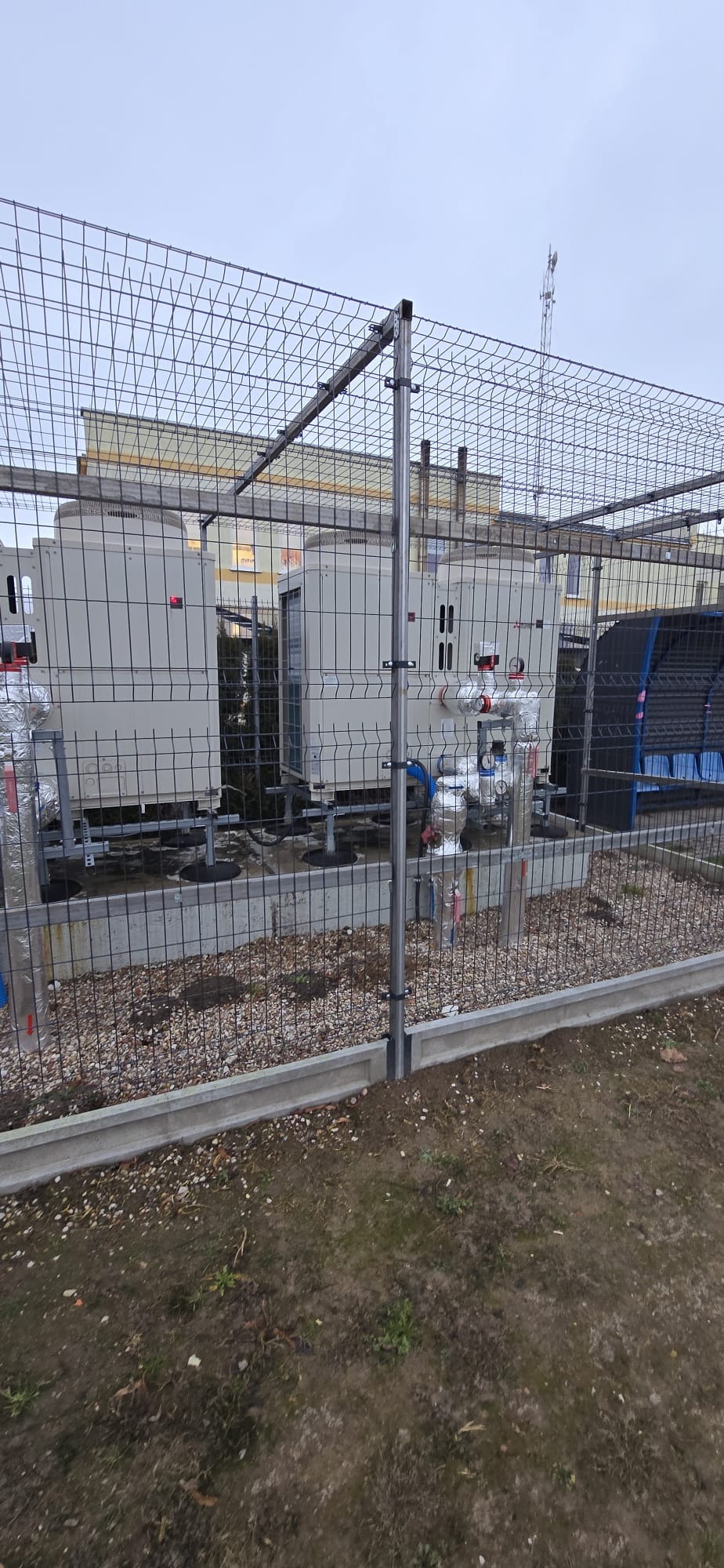 Zewnętrzna instalacja przemysłowa z dwoma agregatami, rurami i zaworami, otoczona metalową siatką ochronną. Widoczne elementy mocowania i podłączenia.