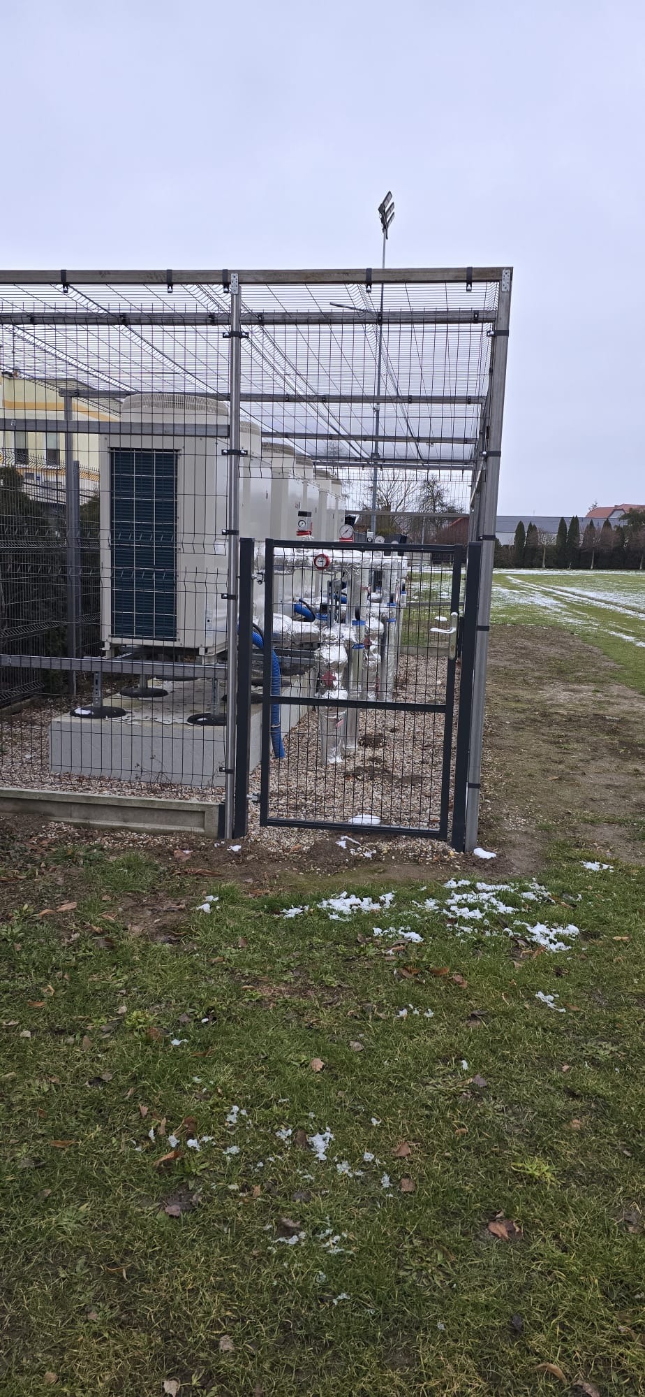 Zabezpieczona instalacja pomp ciepła z rzędami urządzeń za metalową siatką i bramką, na zewnątrz w pochmurny dzień. Widoczne elementy instalacji i teren.
