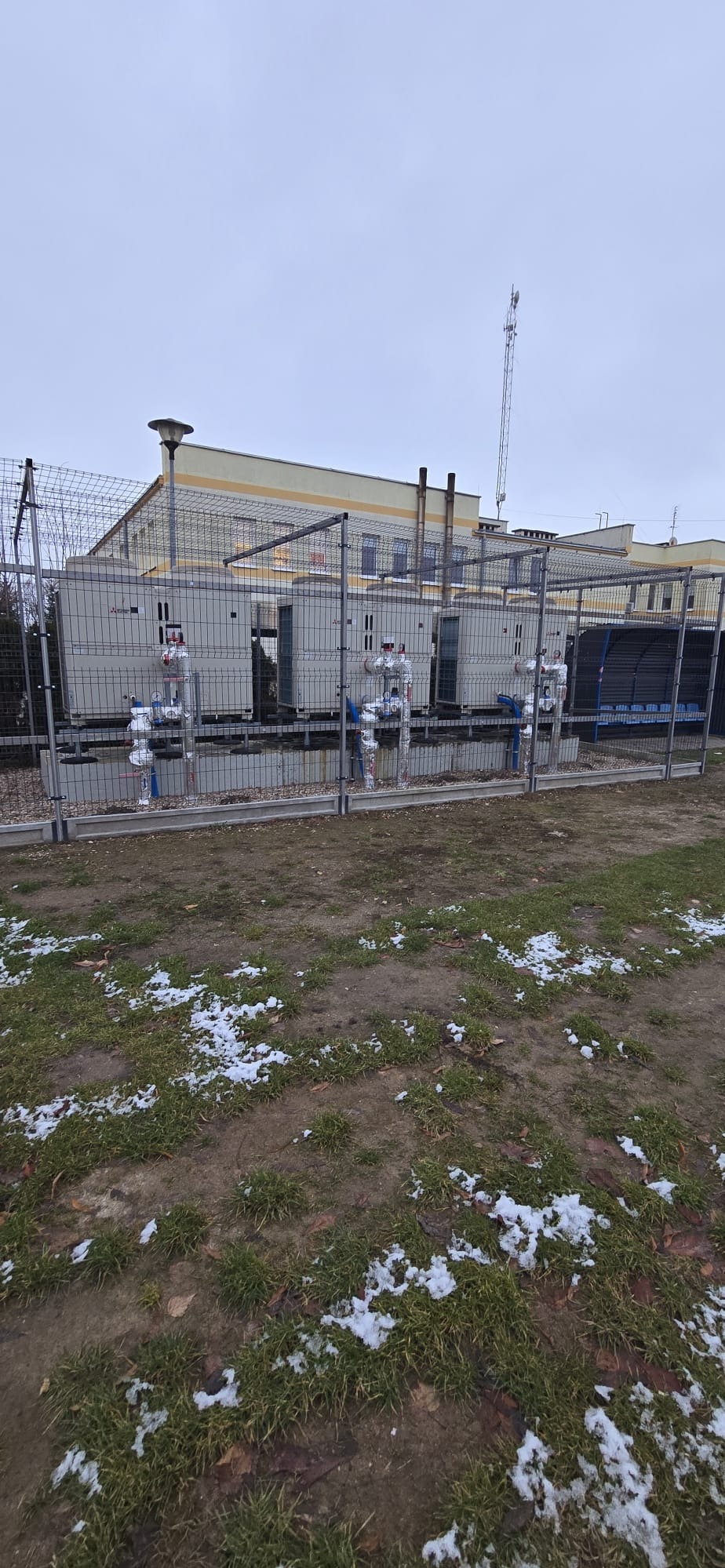 Instalacja kilku pomp ciepła na zewnątrz budynku, zabezpieczona ogrodzeniem z siatki. Widoczne rury i elementy instalacji. Pochmurne niebo w tle.