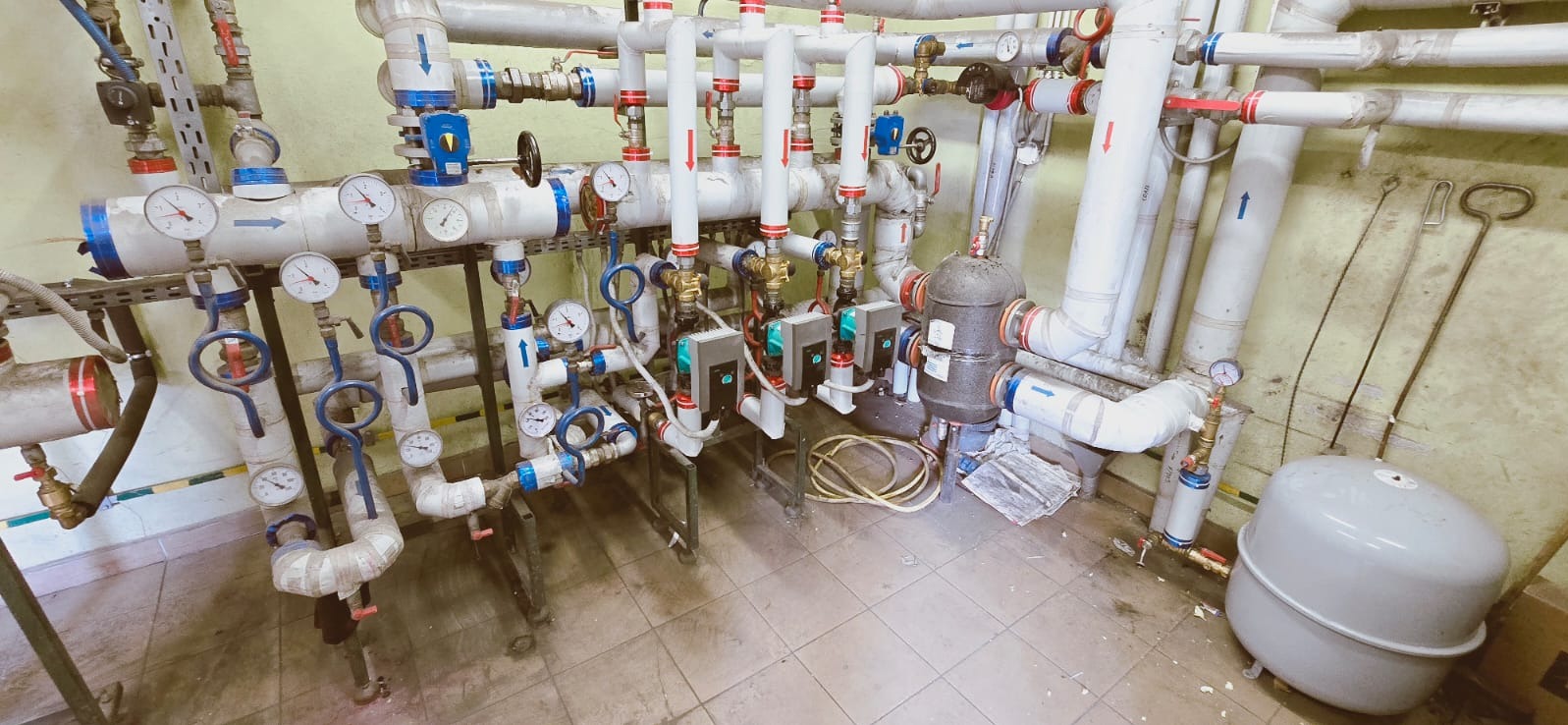 Rozbudowana instalacja grzewcza z rurami, zaworami i manometrami w pomieszczeniu technicznym. Widoczne pompy obiegowe i zbiornik wyrównawczy.